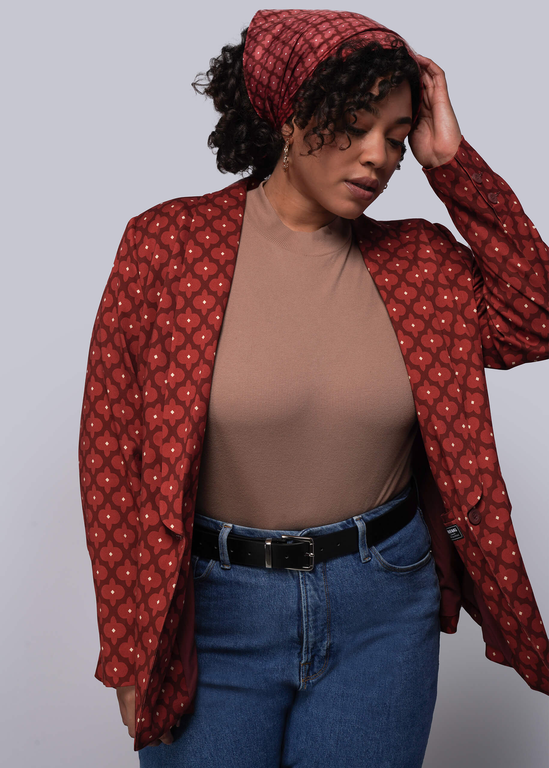 Person wearing a red patterned jacket over a brown top and blue jeans on a gray background