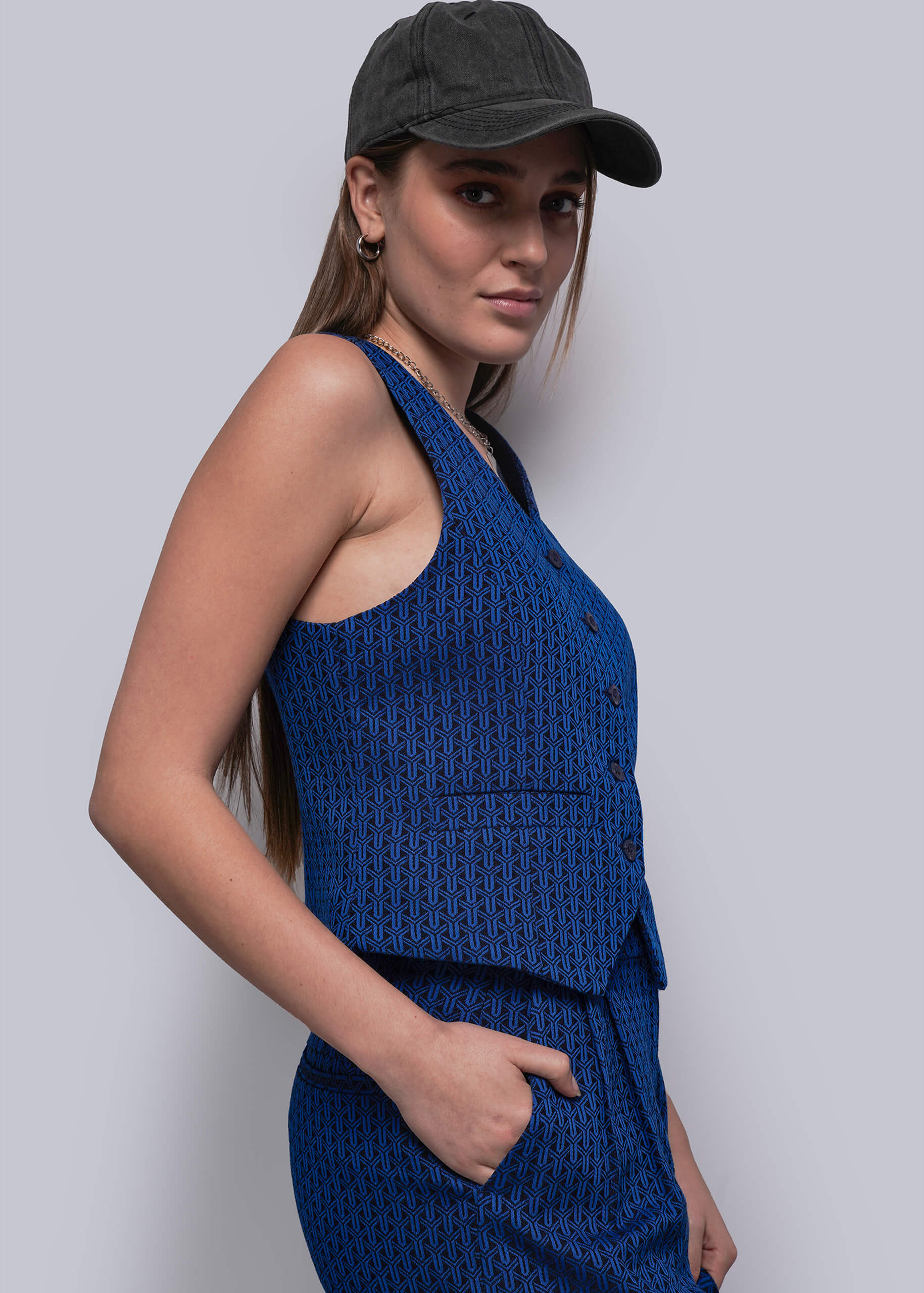 Woman wearing a blue vest and pants with a black cap against a light gray background