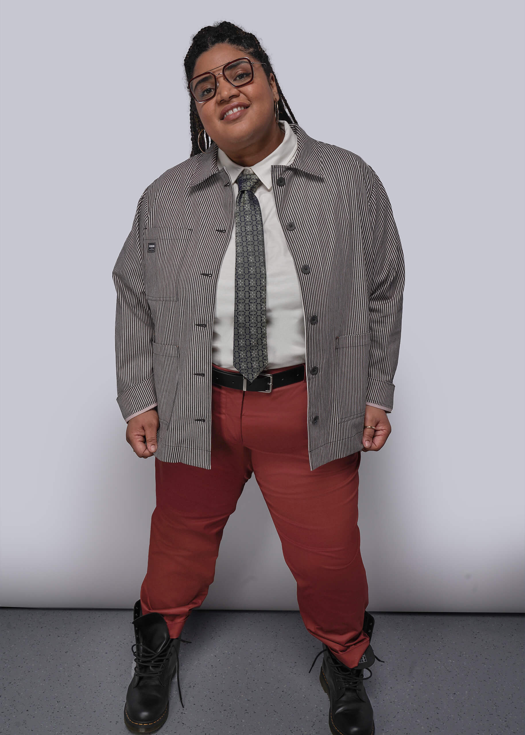 Person wearing a gray jacket, white shirt, red pants, and black shoes against a plain background
