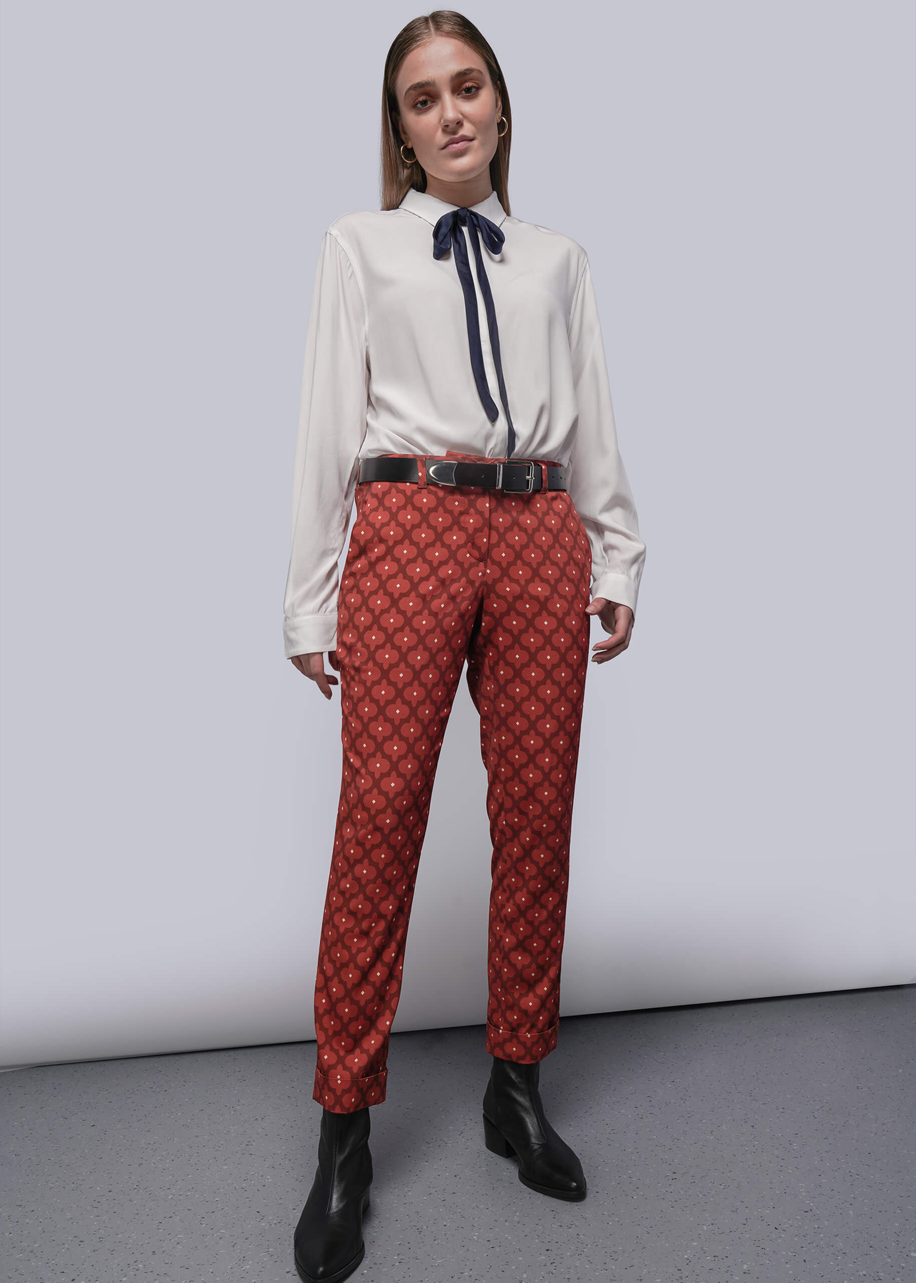 S=Woman wearing a white blouse with a navy tie and red patterned pants against a plain background