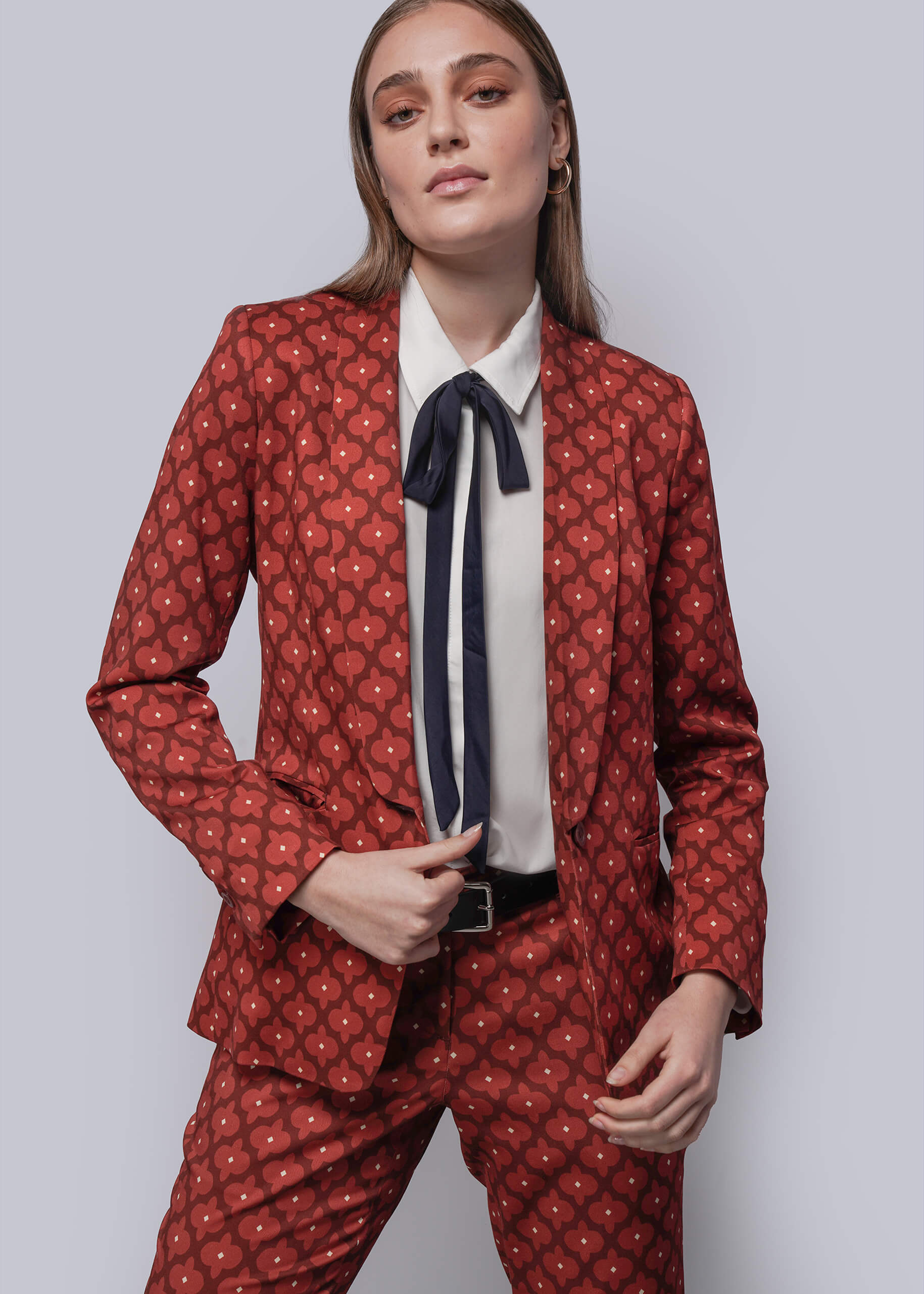 S=Woman wearing a red patterned suit with a white shirt with navy bow on a light gray background