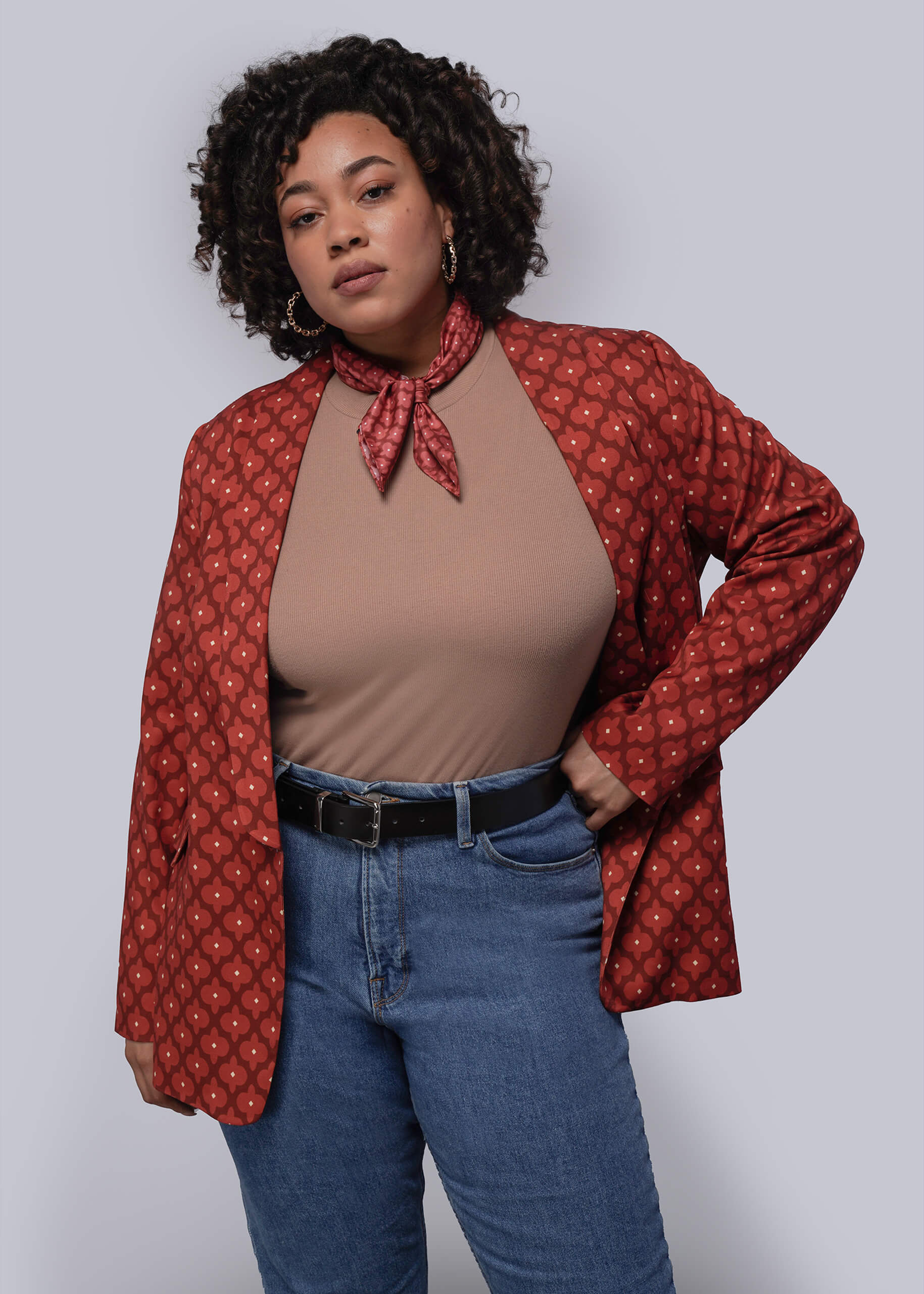 1X=Woman wearing a red patterned jacket over a brown top and blue jeans on a plain background