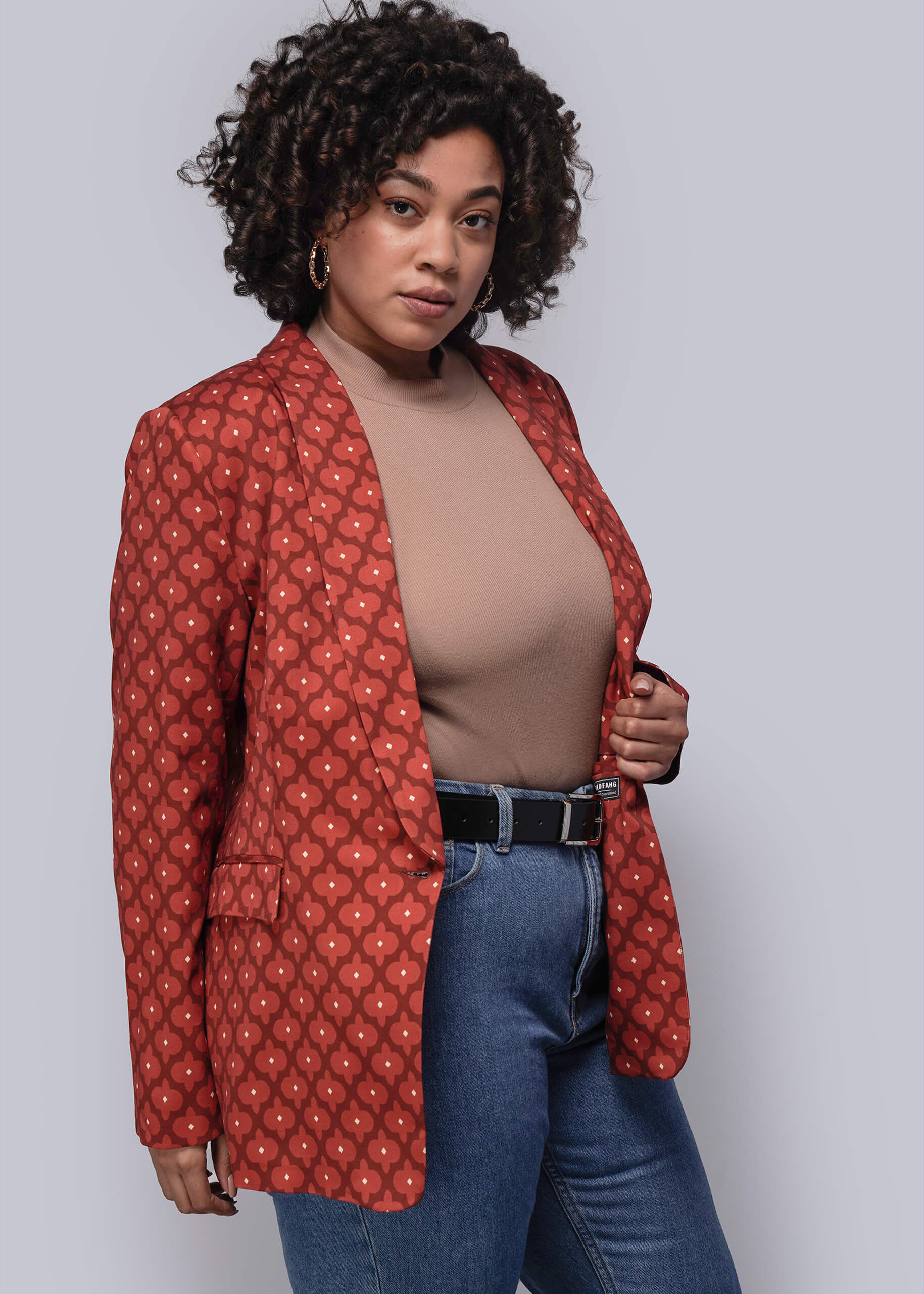 1X=Woman wearing a red patterned blazer over a beige top and blue jeans on a plain background