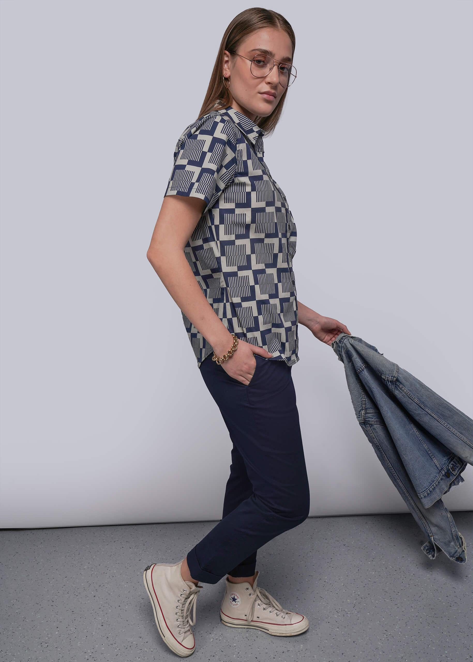 Person wearing a patterned shirt and dark pants, holding a denim jacket against a plain background