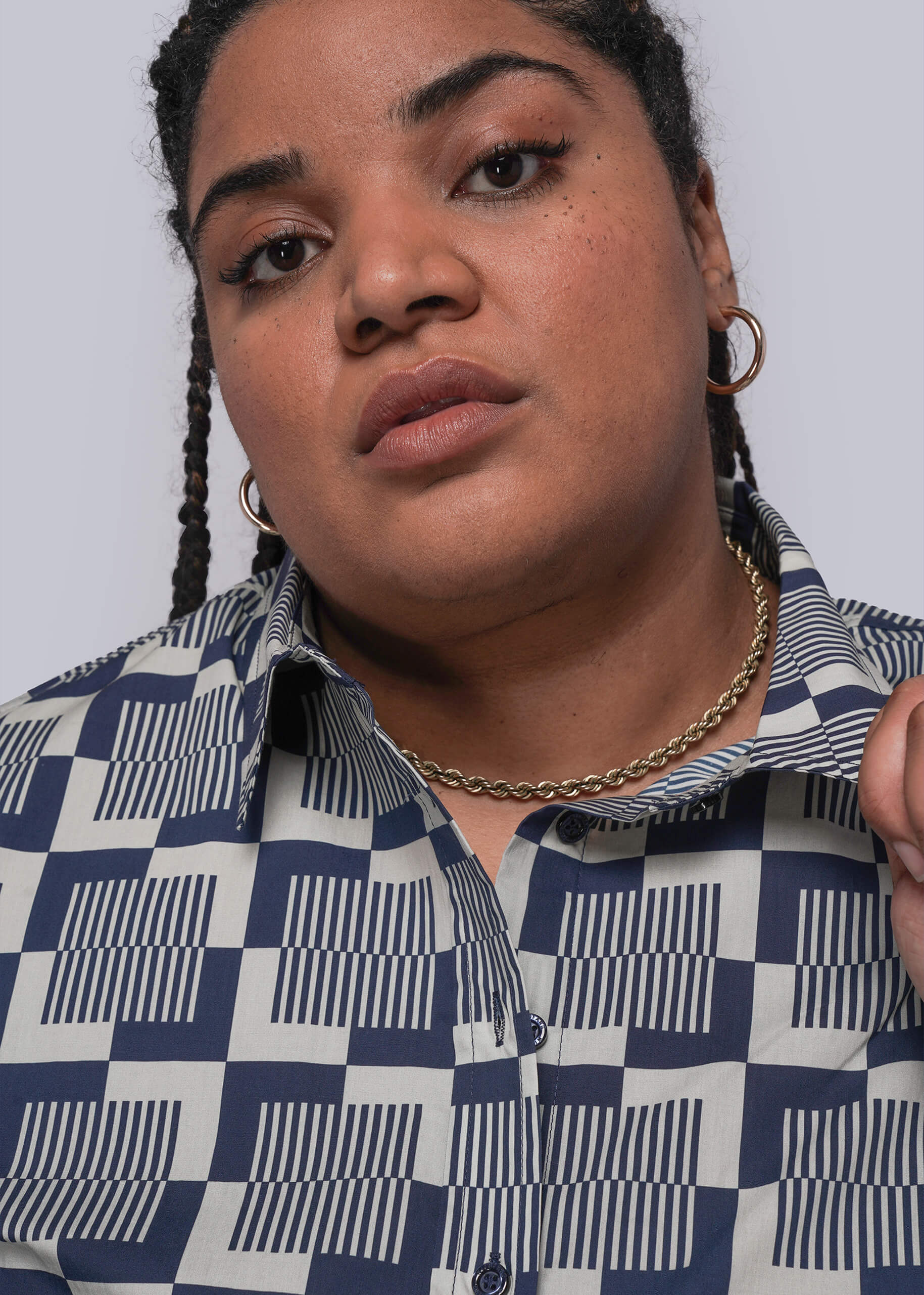 1X=Person wearing a blue and white checkered shirt with braided hair against a plain background