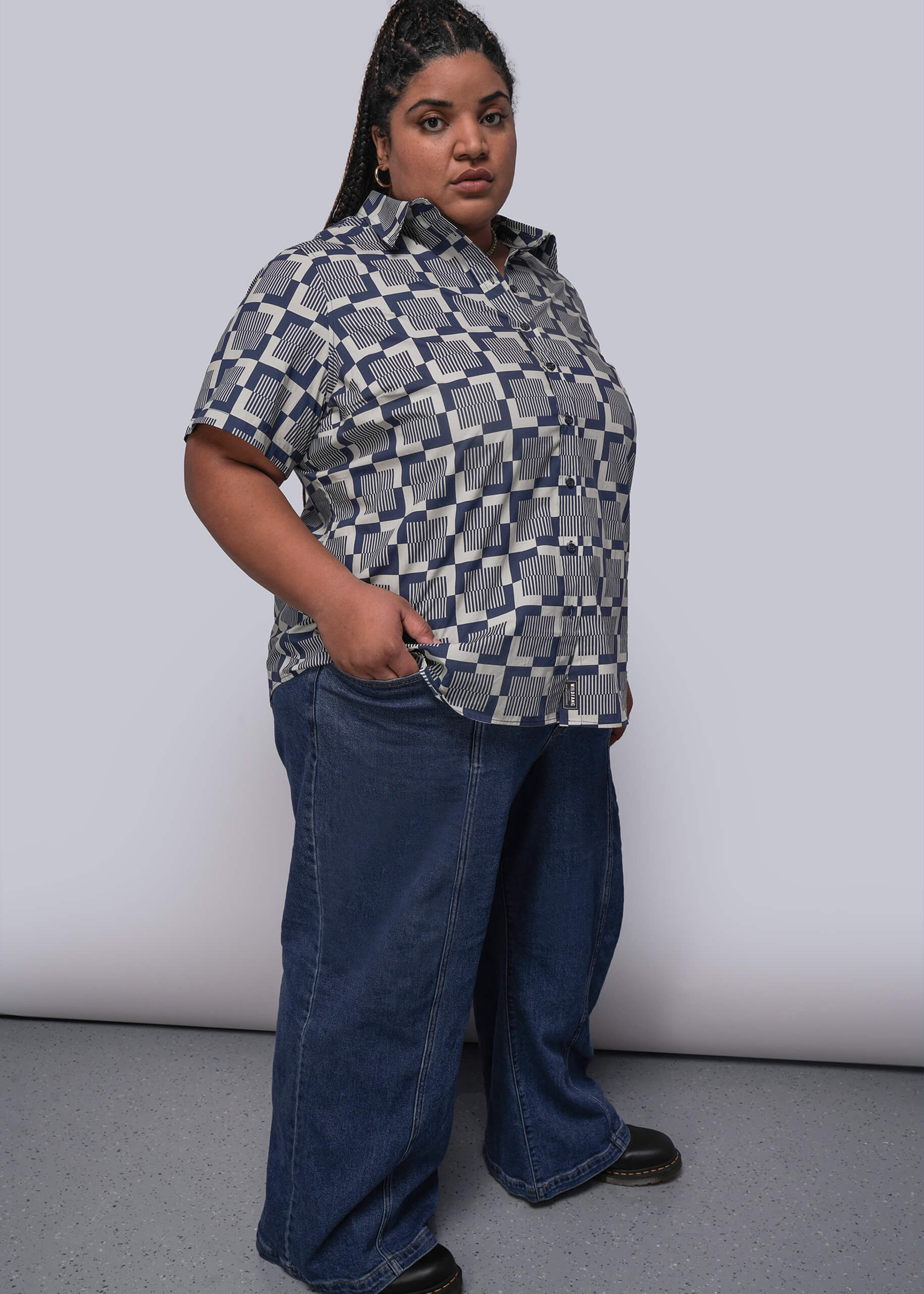 1X=Person wearing a patterned shirt and blue jeans against a plain background