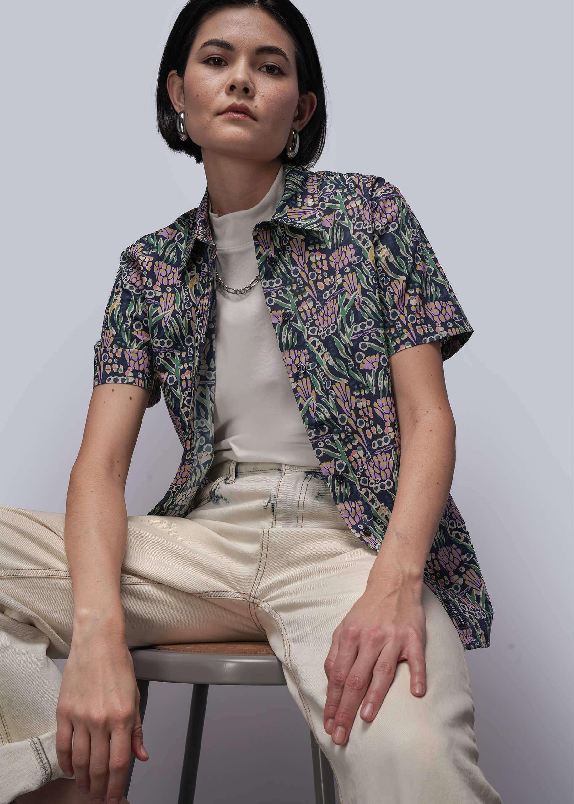 S=Woman sitting on a chair wearing a floral shirt over a white top and beige pants.