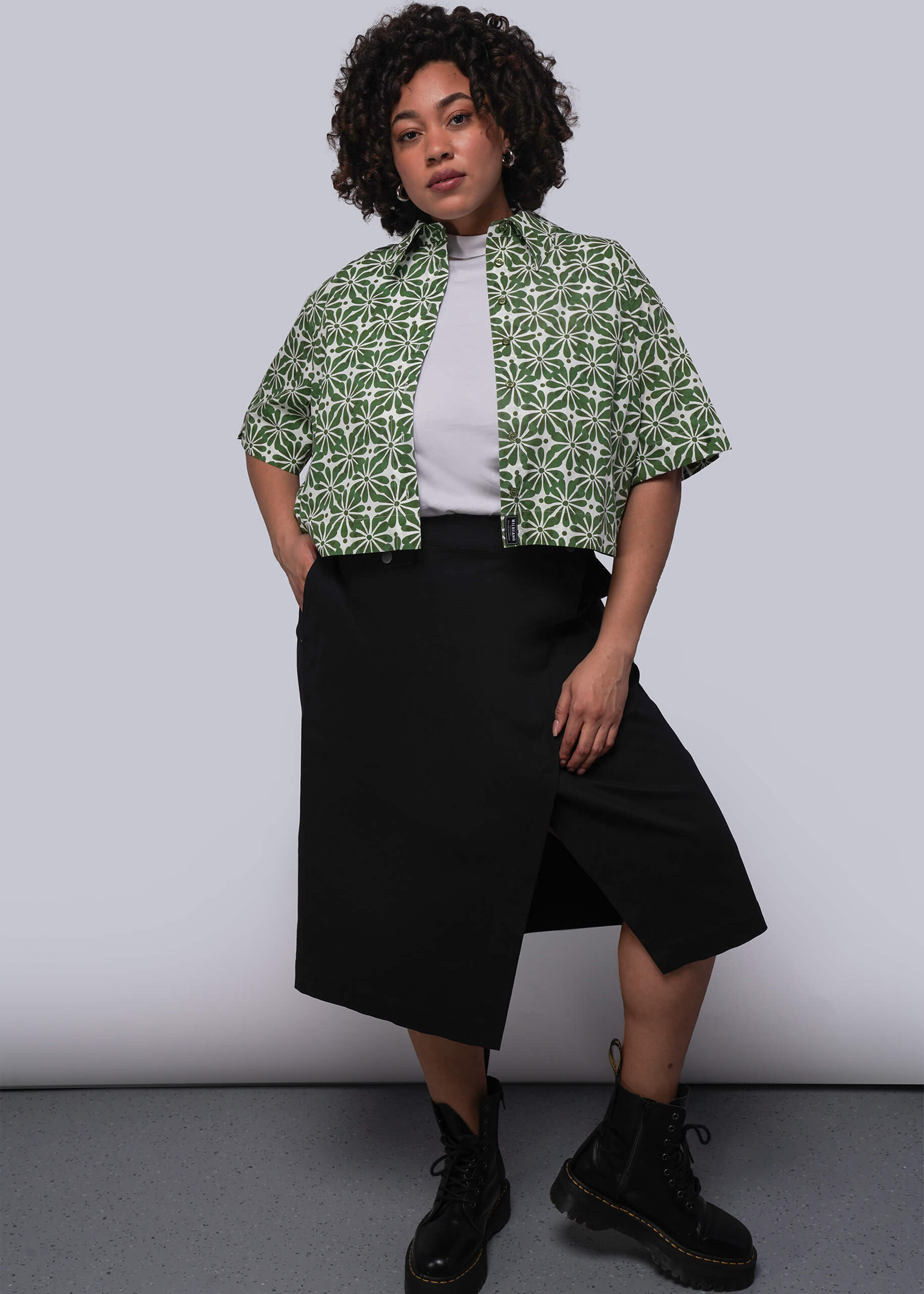 1X=Person wearing a green patterned cropped button up, white shirt, black skirt, and black shoes on a plain background
