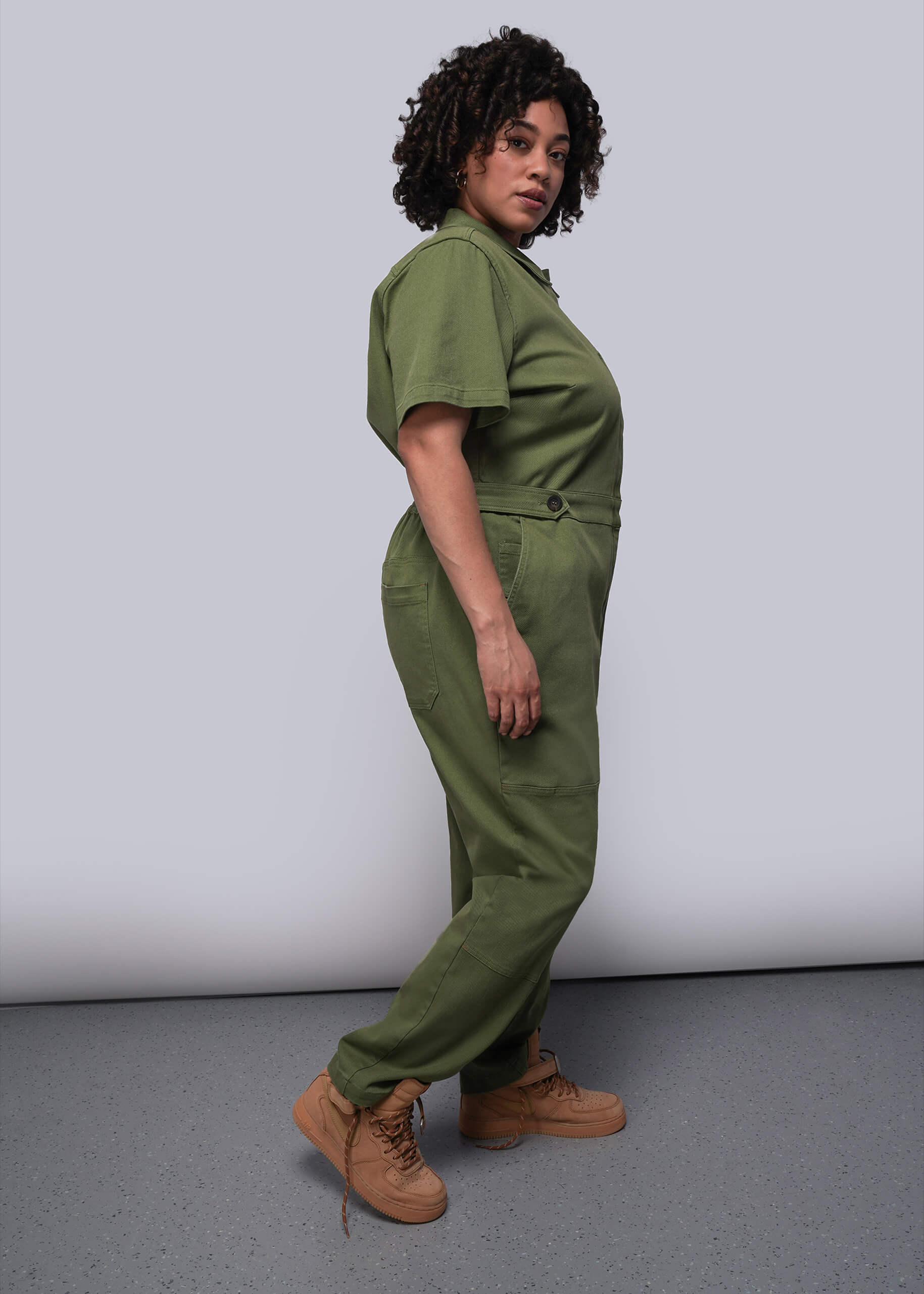 1X=Person wearing a green jumpsuit standing against a plain background