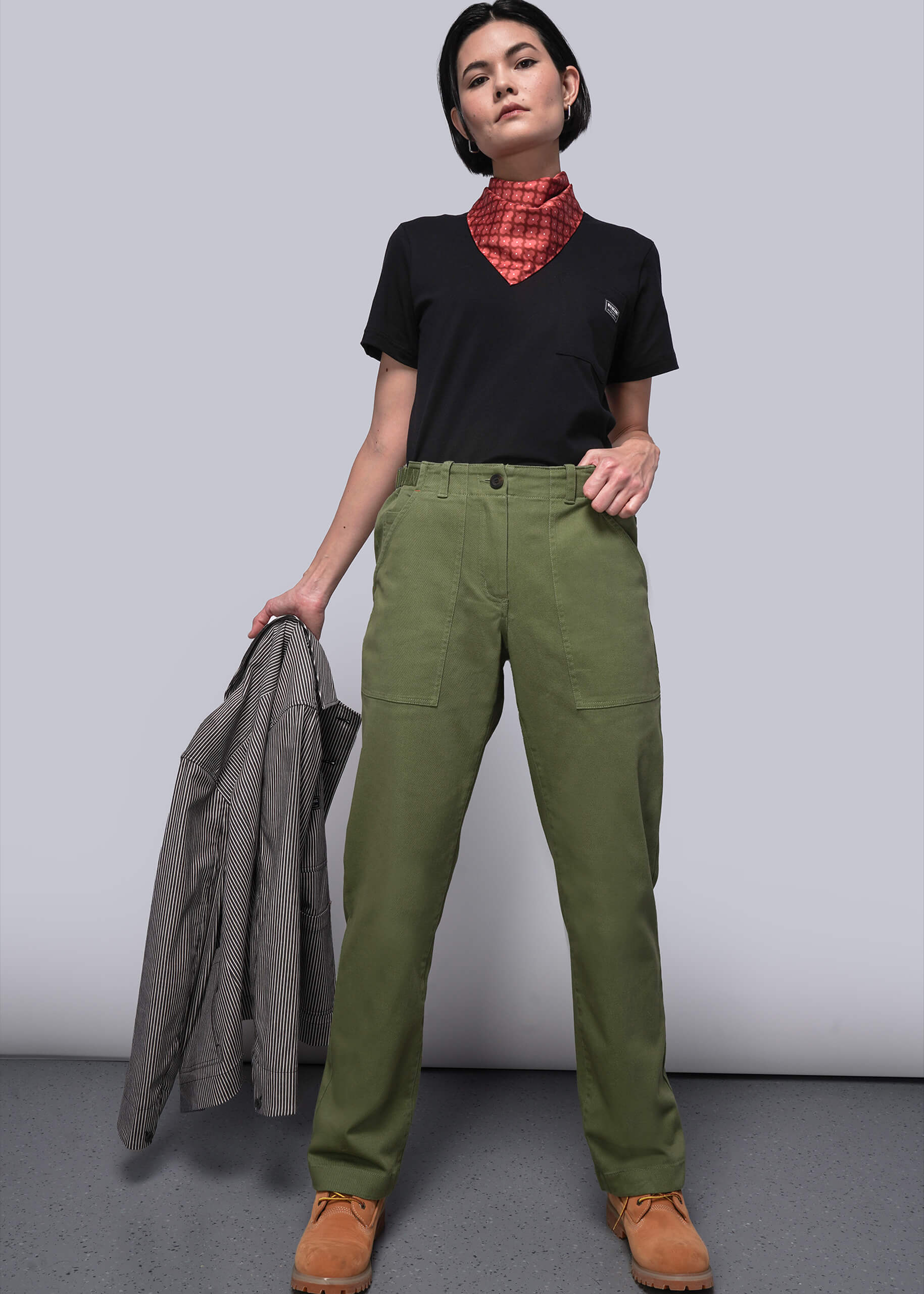 S=Person wearing green pants and a black shirt with a red scarf, holding a gray jacket against a plain background.