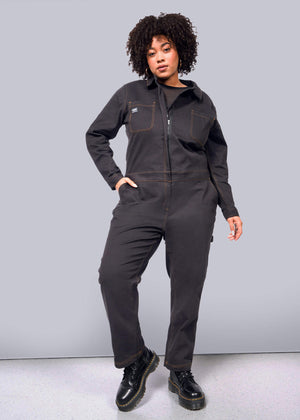 1X=A person with curly hair confidently stands in The Essential Long Sleeve Coverall, made of brushed stretch cotton, while wearing black boots. One hand in the pocket and the other on their hip against a plain white background.