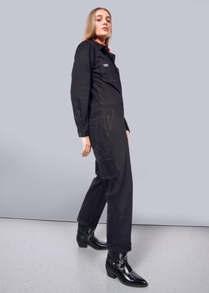 S=Against a white wall, the person exudes confidence in The Essential Long Sleeve Coverall and shiny boots. Long hair frames their face as they gaze into the camera, embodying a bold style.