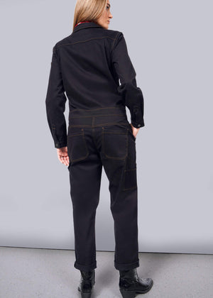 S=A woman with long hair stands facing away, modeling The Essential Long Sleeve Coverall. This oversized brushed stretch cotton jumpsuit, featuring brown stitching and 10 pockets, pairs perfectly with her black boots against a plain white backdrop. Her right hand confidently rests on her hip.