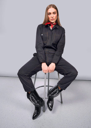 S=A person with long hair sits on a stool against a white background, wearing The Essential Long Sleeve Coverall in an oversized fit. Its crafted from brushed stretch cotton with visible stitching and pairs perfectly with their red bandana and black pointed cowboy boots, hands relaxed on their knees.