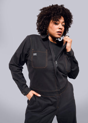 1X=A person with curly hair confidently rocks The Essential Long Sleeve Coverall, an oversized black jumpsuit crafted from brushed stretch cotton with brown stitching. With one hand in their pocket and the other adjusting the collar, they stand poised against a plain white background.