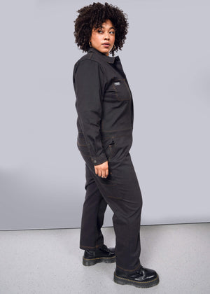 1X=A person with curly hair stands sideways, gazing at the camera in a black Essential Long Sleeve Coverall made from brushed stretch cotton. It has an oversized fit and 10 pockets, paired with sleek black boots against a plain white background.