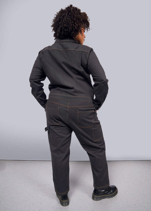 1X=A person with curly hair faces away from the camera, wearing The Essential Long Sleeve Coverall, an oversized black jumpsuit crafted in brushed stretch cotton. They match it with black shoes atop a light gray floor, all set against a plain white background, exuding effortless style and function.