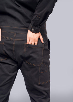 1X=A person in The Essential Long Sleeve Coverall, featuring black fabric with orange stitching and made from brushed stretch cotton, is shown from the back on a white background. The oversized fit allows one hand to be partially tucked into a back pocket.