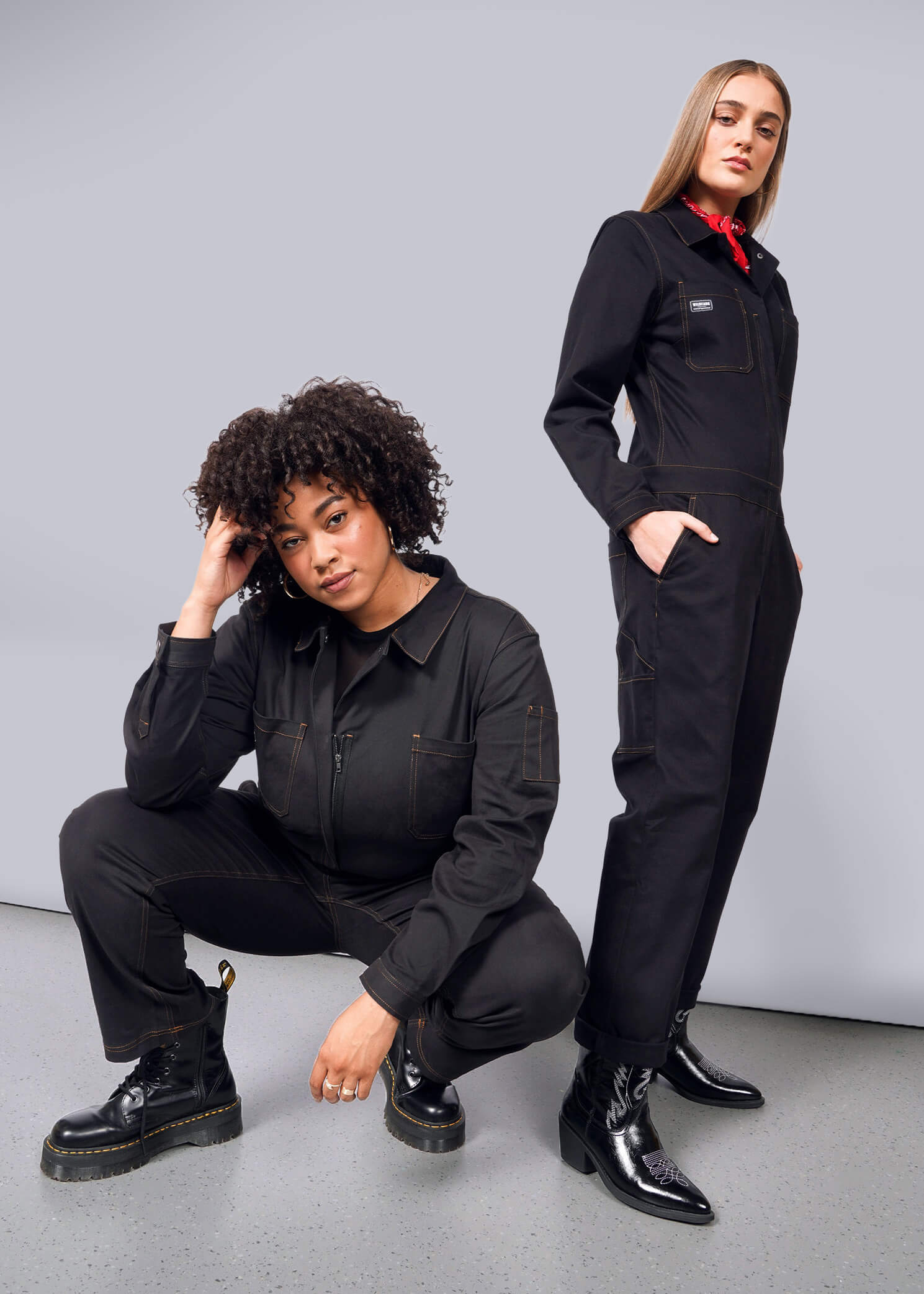 Two women wearing The Essential Long Sleeve Coverall in black pose against a white backdrop. One crouches with curly hair, while the other stands confidently with long, straight hair and a red bandana, both showcasing an oversized fit in brushed stretch cotton.