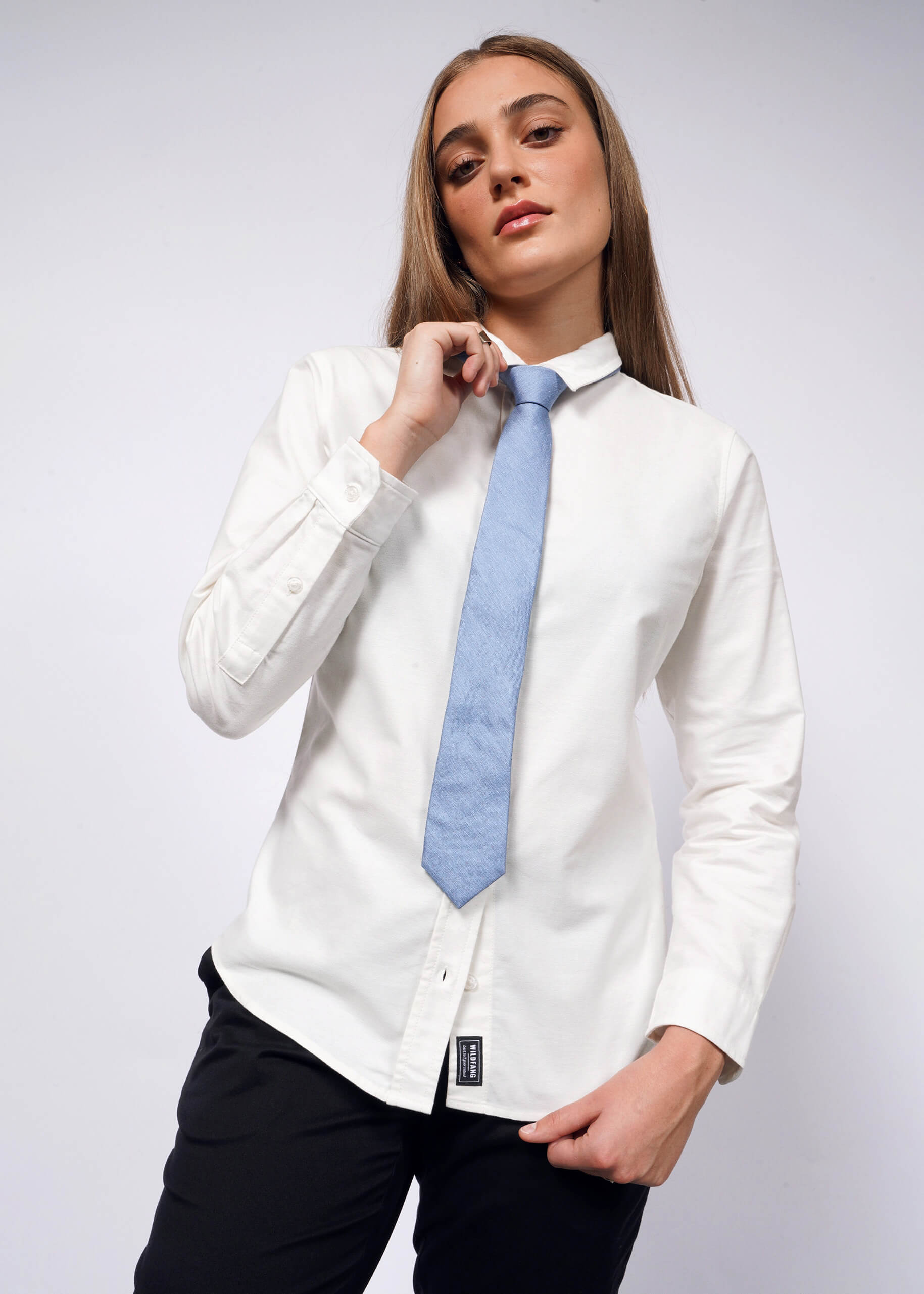 Two individuals pose confidently against a white background. One sports a tailored fit with The Essential Long Sleeve Oxford Button Up, black pants, blue tie, and black Converse sneakers. The other wears a 100% cotton shirt and relaxed blue jeans. Both exude a casual, stylish vibe.