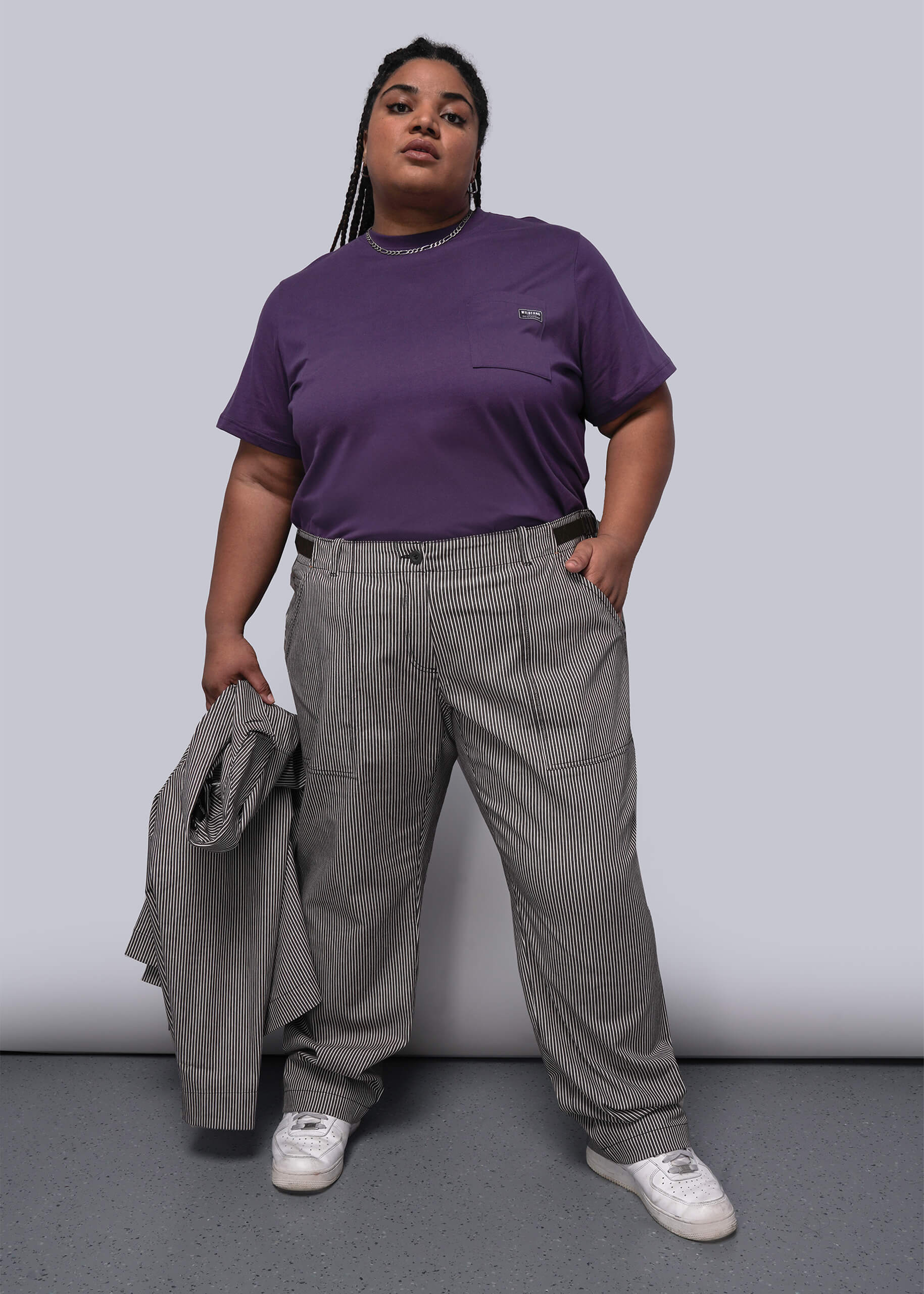 1X=Person wearing a purple t-shirt and striped pants holding a gray jacket against a plain background