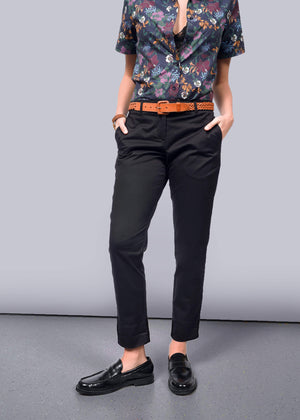 S= A person wearing The Essential Trouser in black, featuring a relaxed mid-rise fit, pairs it with a floral shirt and brown belt. Hands are tucked into pockets, completed with black loafers. The stylish outfit is presented on a light gray floor against a pristine white background.