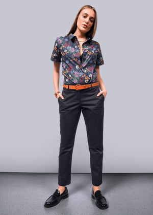 S=The woman stands confidently in a floral short-sleeve shirt and The Essential Trouser in relaxed mid-rise black. Black loafers and a brown belt with a bracelet complete the look. Her hands rest in her pockets against a plain light gray wall and floor, highlighting her poised stance.