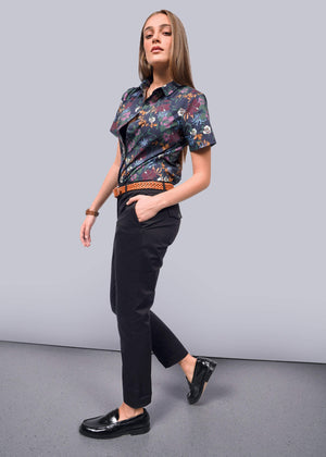 S=In a bright, minimalist setting, a person with long hair confidently strides wearing a short-sleeved patterned shirt and The Essential Trouser. They showcase a patterned belt and shiny loafers, their relaxed mid-rise fit enhancing the effortlessly stylish ensemble.