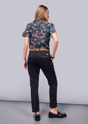 S=A person with long hair stands facing away, showcasing a floral short-sleeve shirt and The Essential Trouser in black with matching shoes. They stand on a light gray floor against a plain white background.