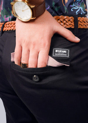 S=A close-up shows someone in The Essential Trouser, featuring a relaxed mid-rise fit and dark finish, accented by a woven brown belt. A WILDFANG tag is visible as their hand rests in a pocket lined with red and white stripes. They wear a watch with a brown leather strap and a ring for a timeless look.