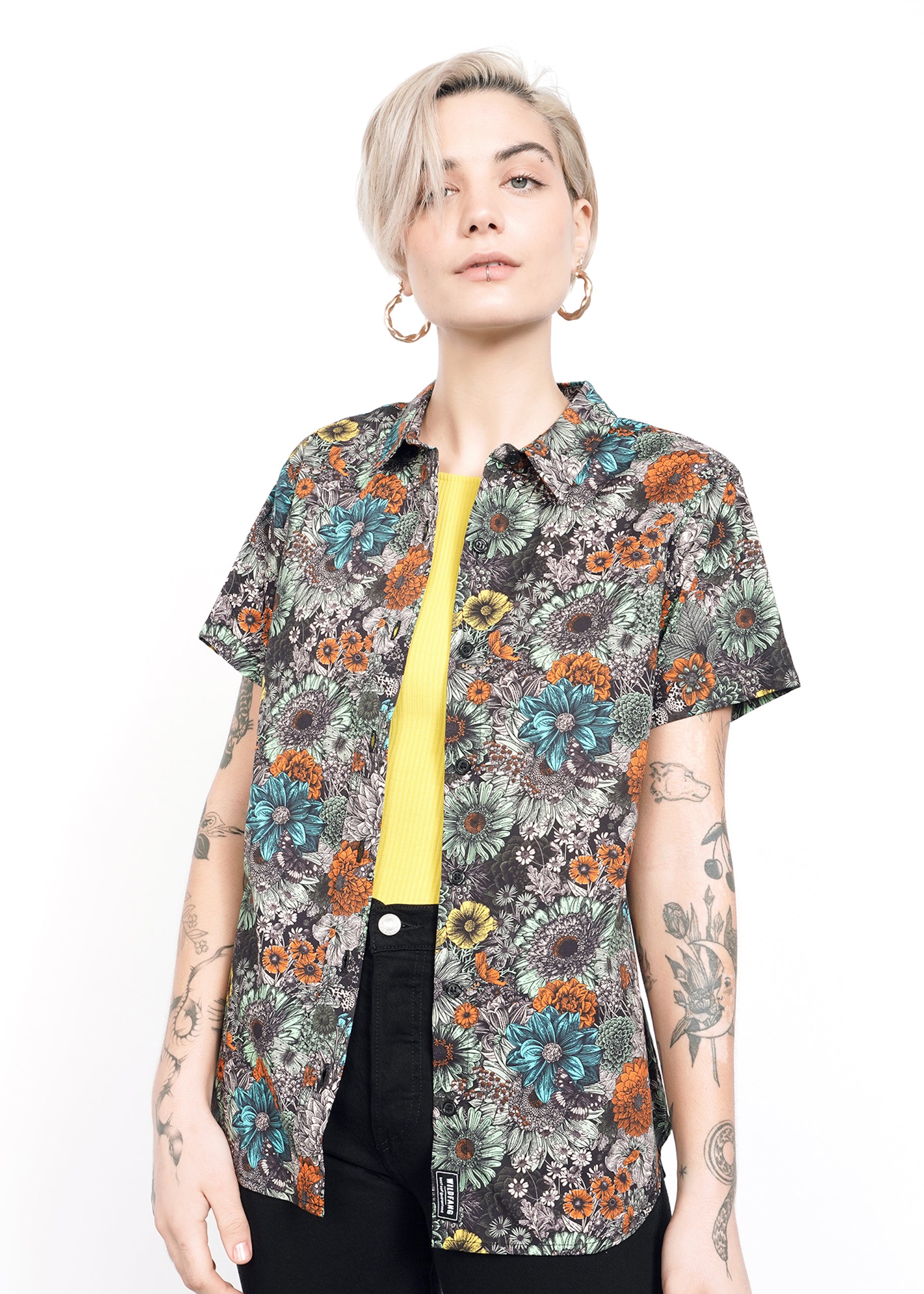 S= A person with short blonde hair stands confidently against a white background, wearing The Essential Button Up—a tailored-fit, floral-patterned shirt made of 100% cotton—with a yellow top and black pants. They have tattoos on both arms and are wearing hoop earrings.