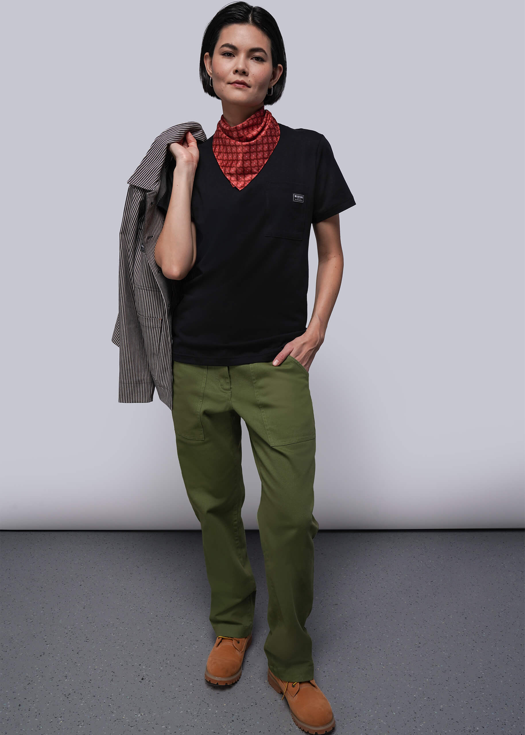 S=Person wearing a black shirt, green pants, and brown shoes against a plain background