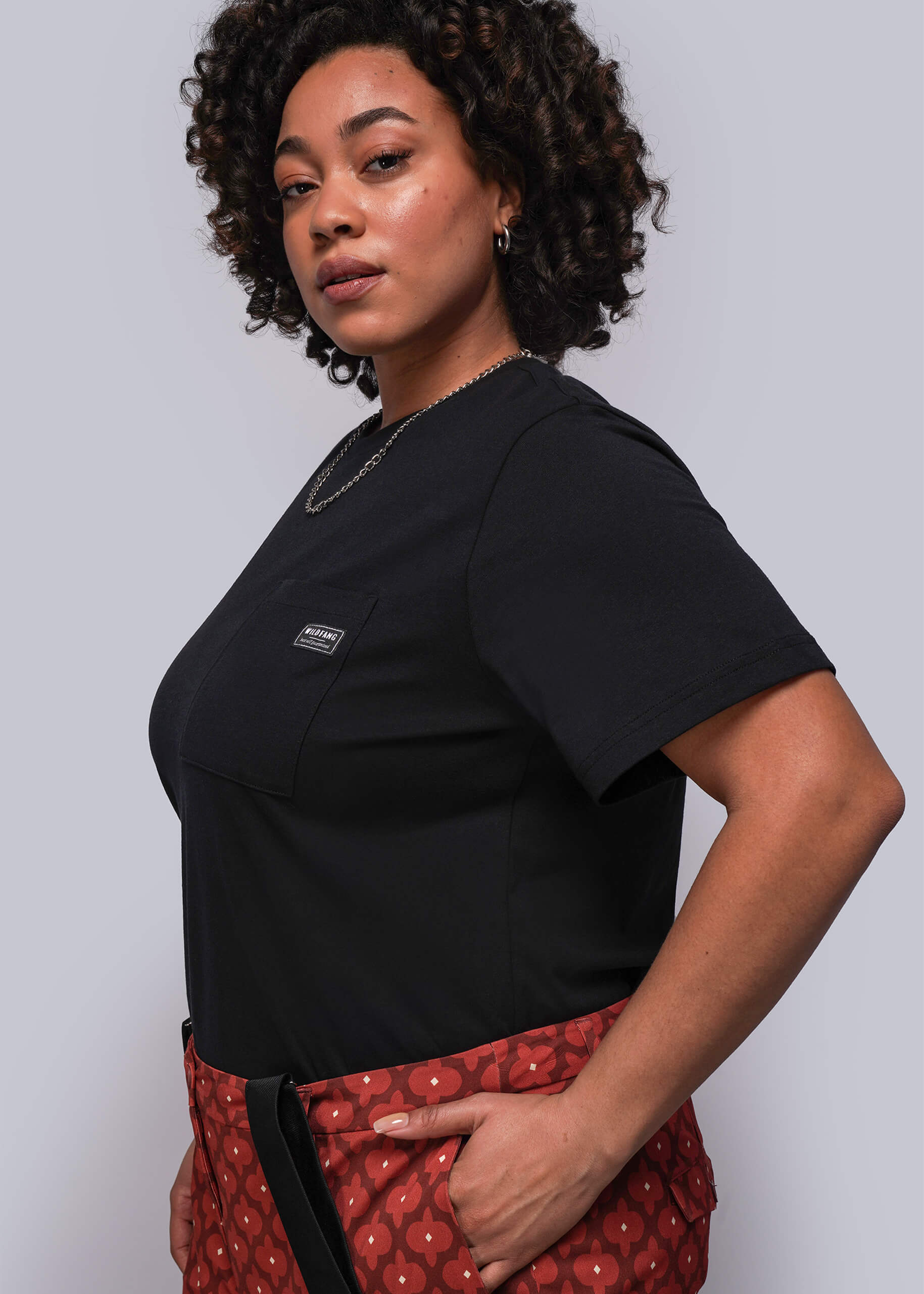 1X=Woman wearing a black top with a logo and red patterned pants against a plain background