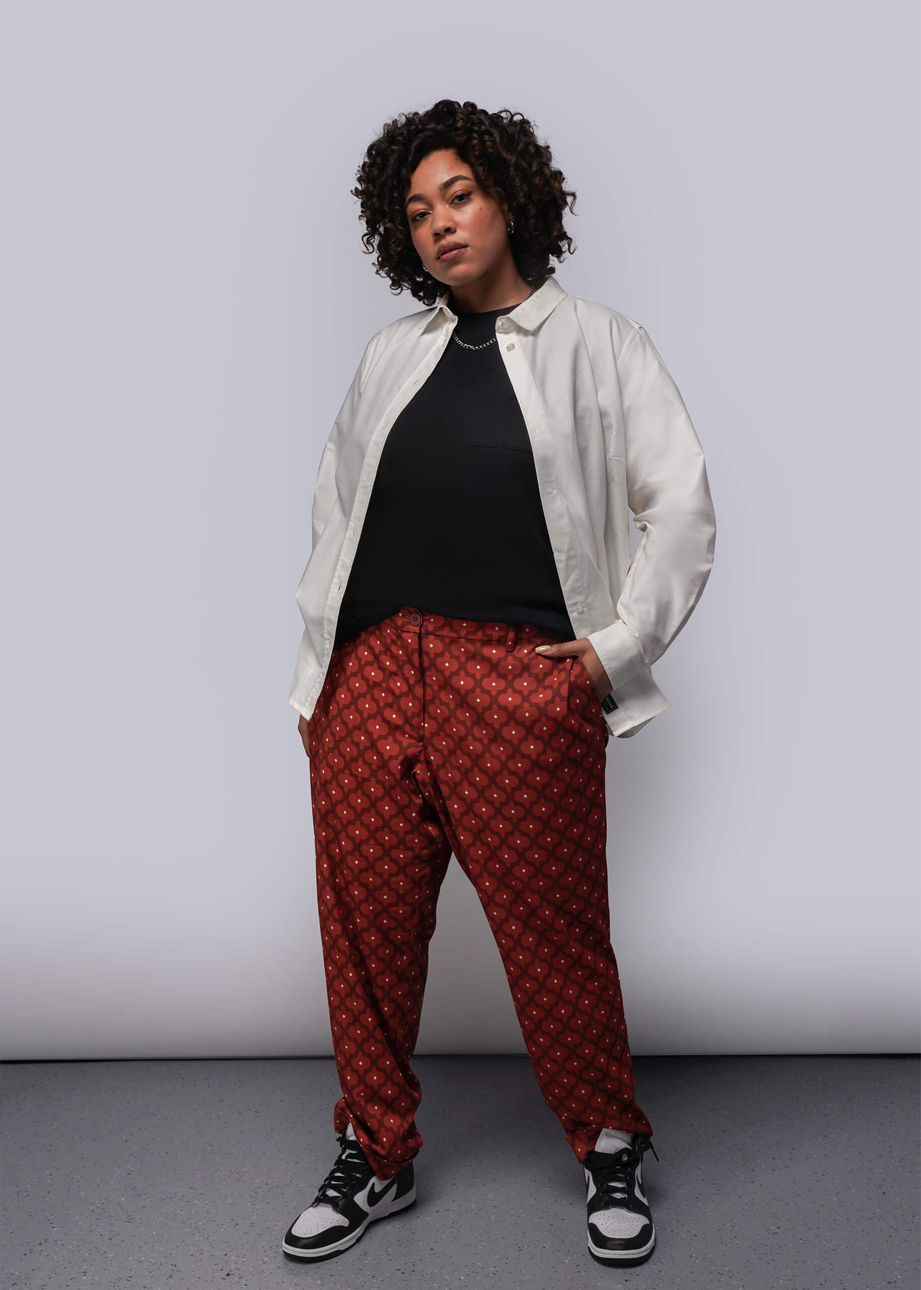 1X=Person wearing a white shirt, black top, and red patterned pants against a plain background