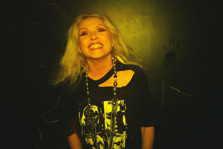 Person with long blonde hair wearing a black top with graphic design against a dark background