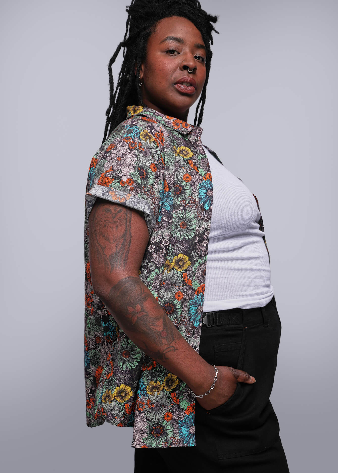 1X=Person wearing a colorful floral shirt over a white top with black pants on a gray background