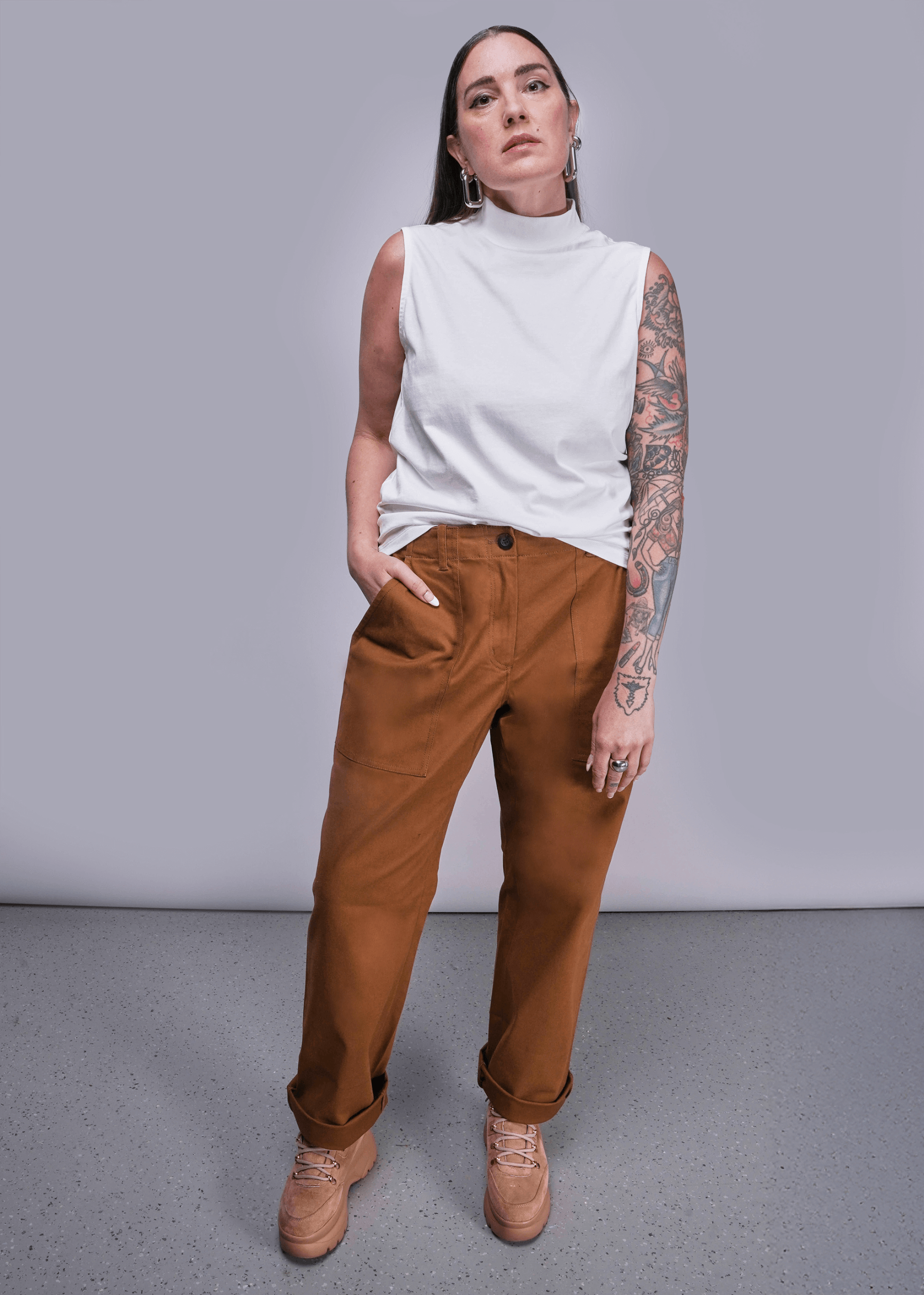 Model with long brown hair stands in front of light grey background wearing The Everyday Mock Neck Tank in White and The Essential Utility Pant in Walnut.