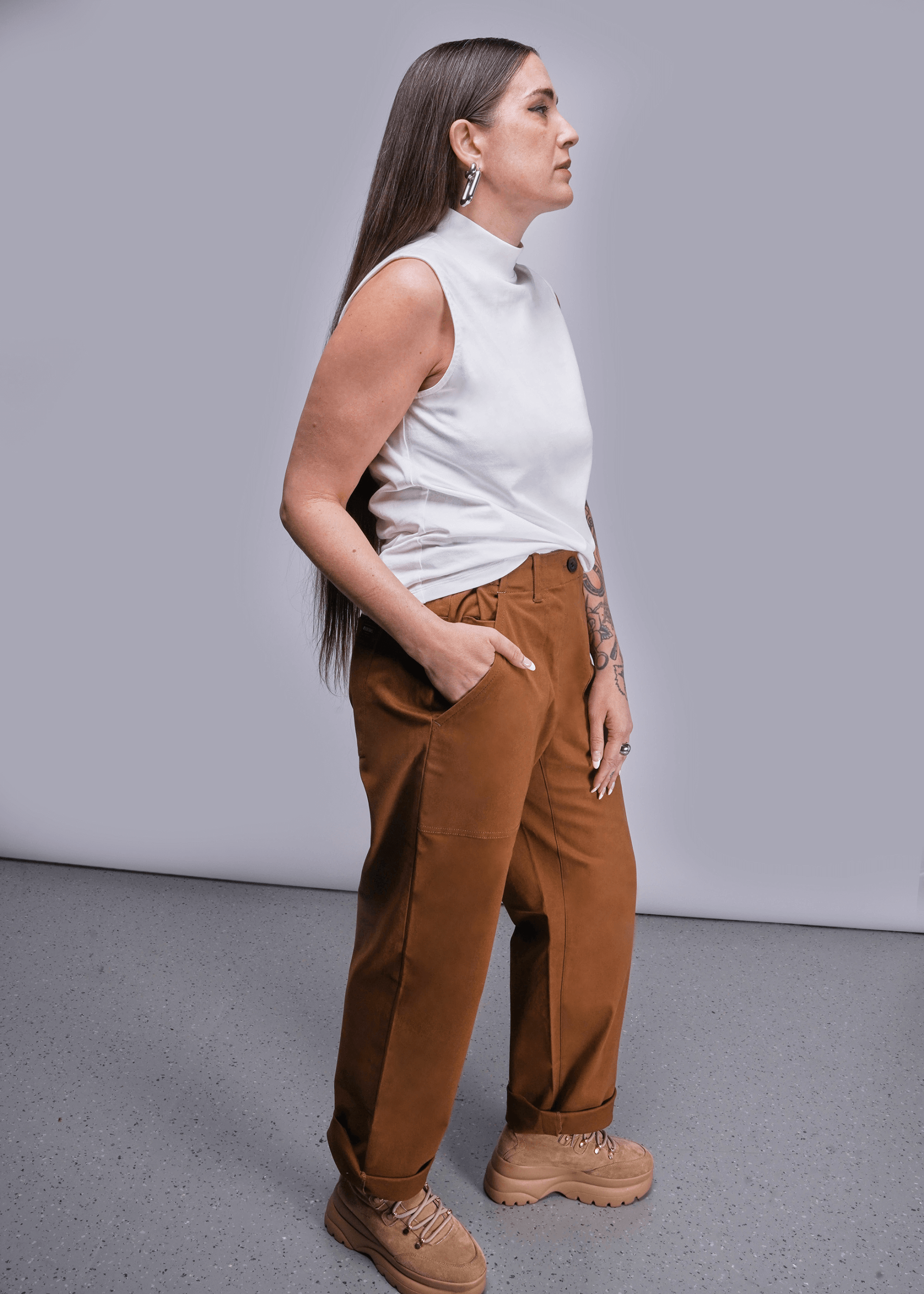 Side view of model with long brown hair standing in front of light grey background wearing The Everyday Mock Neck Tank in White and The Essential Utility Pant in Walnut with hand in pocket.