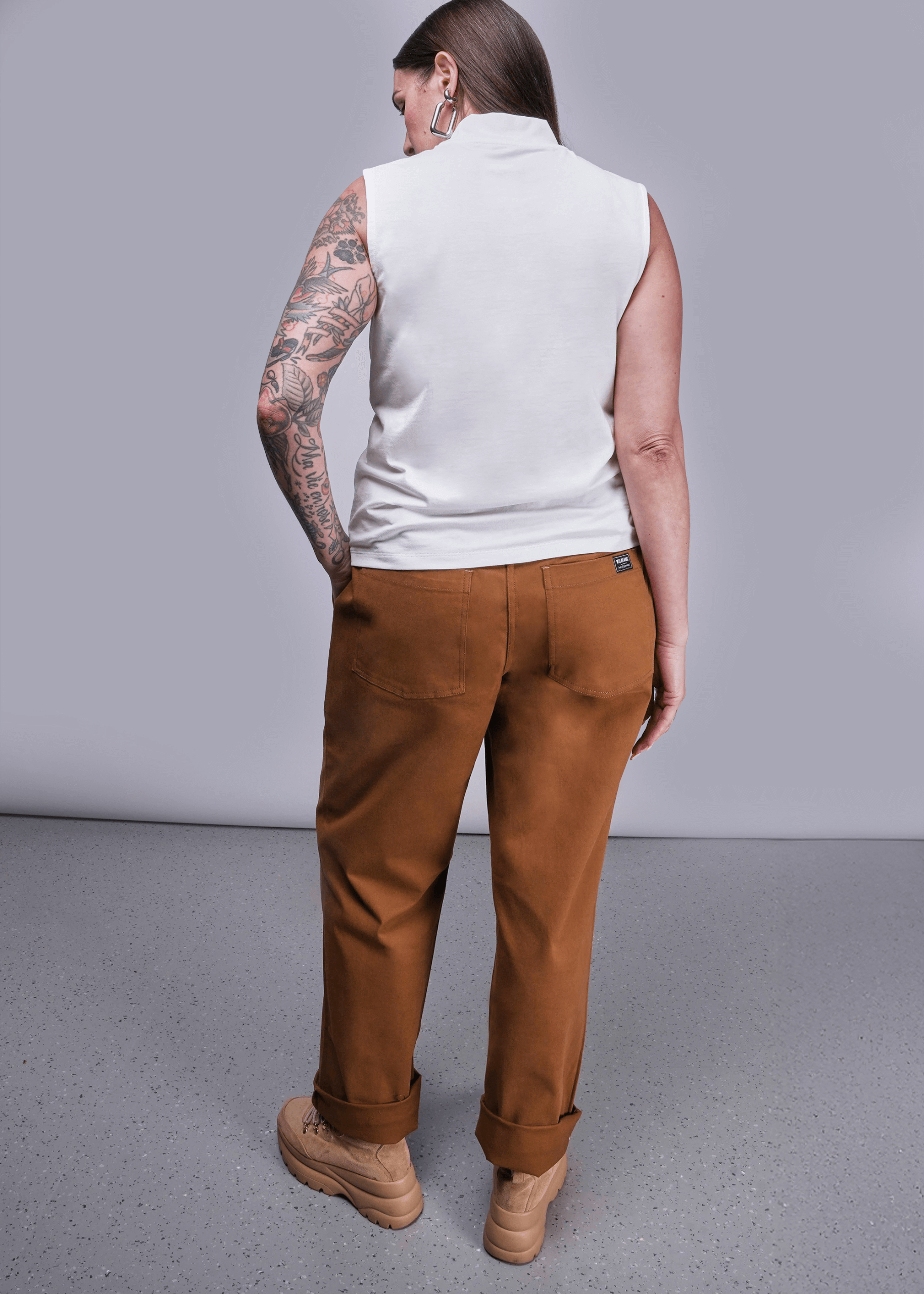 Back view of model with long brown hair standing in front of light grey background wearing The Everyday Mock Neck Tank in White and The Essential Utility Pant in Walnut with hand in pocket.