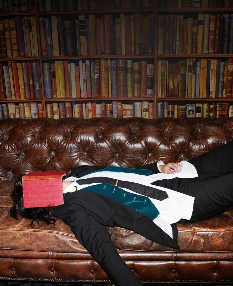 Person wearing black suit lays on leather couch in library with red book over face.