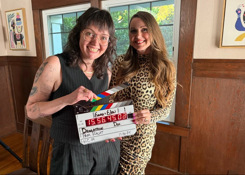 Two women holding a clapperboard in a room with wooden walls and a window.