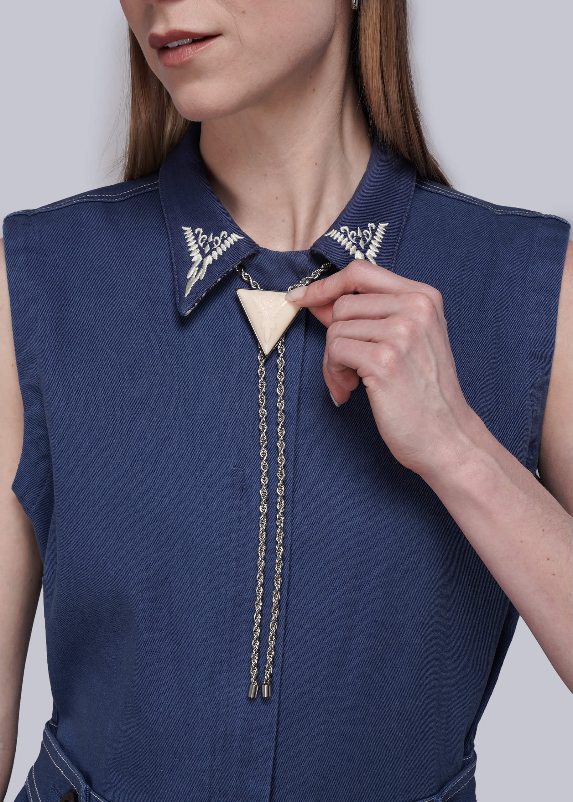 Person wearing denim coverall and holding pendant of silver bolo tie against grey background.