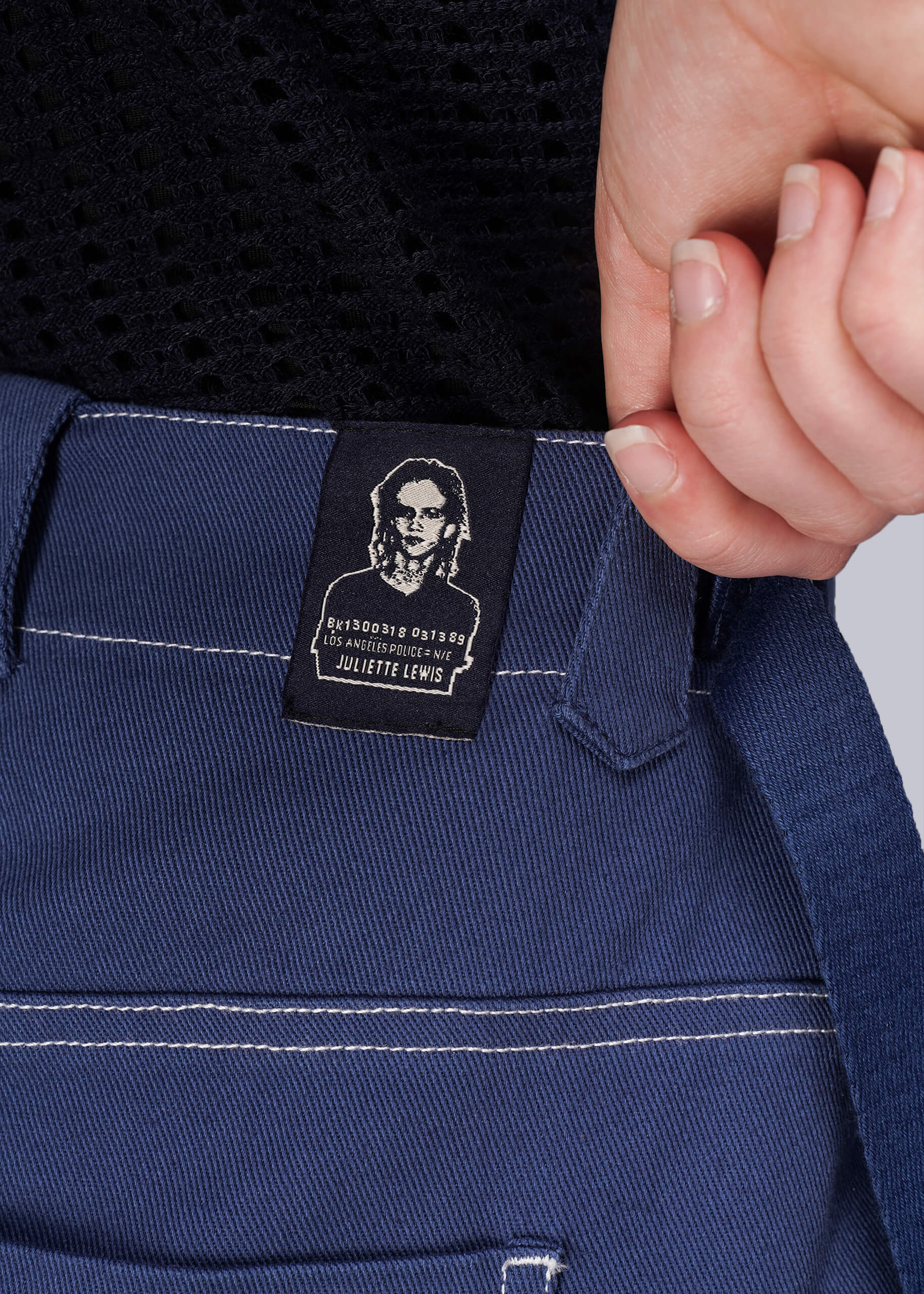 S=Close-up of a blue denim pant showing a label featuring a mugshot of Juliette Lewis.