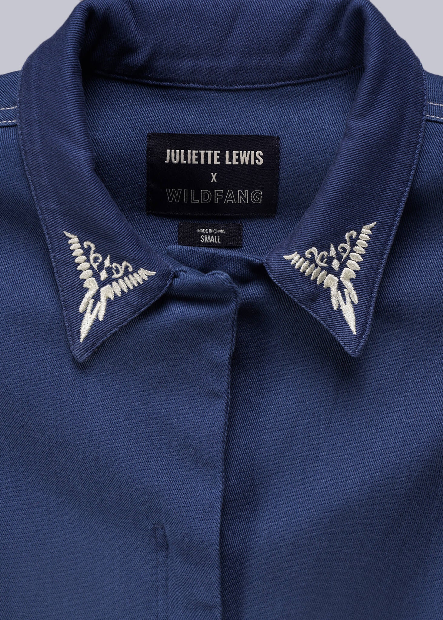 1X=Close up of denim coverall with embroidered collar and 'Juliette Lewis x Wildfang' label on a gray background