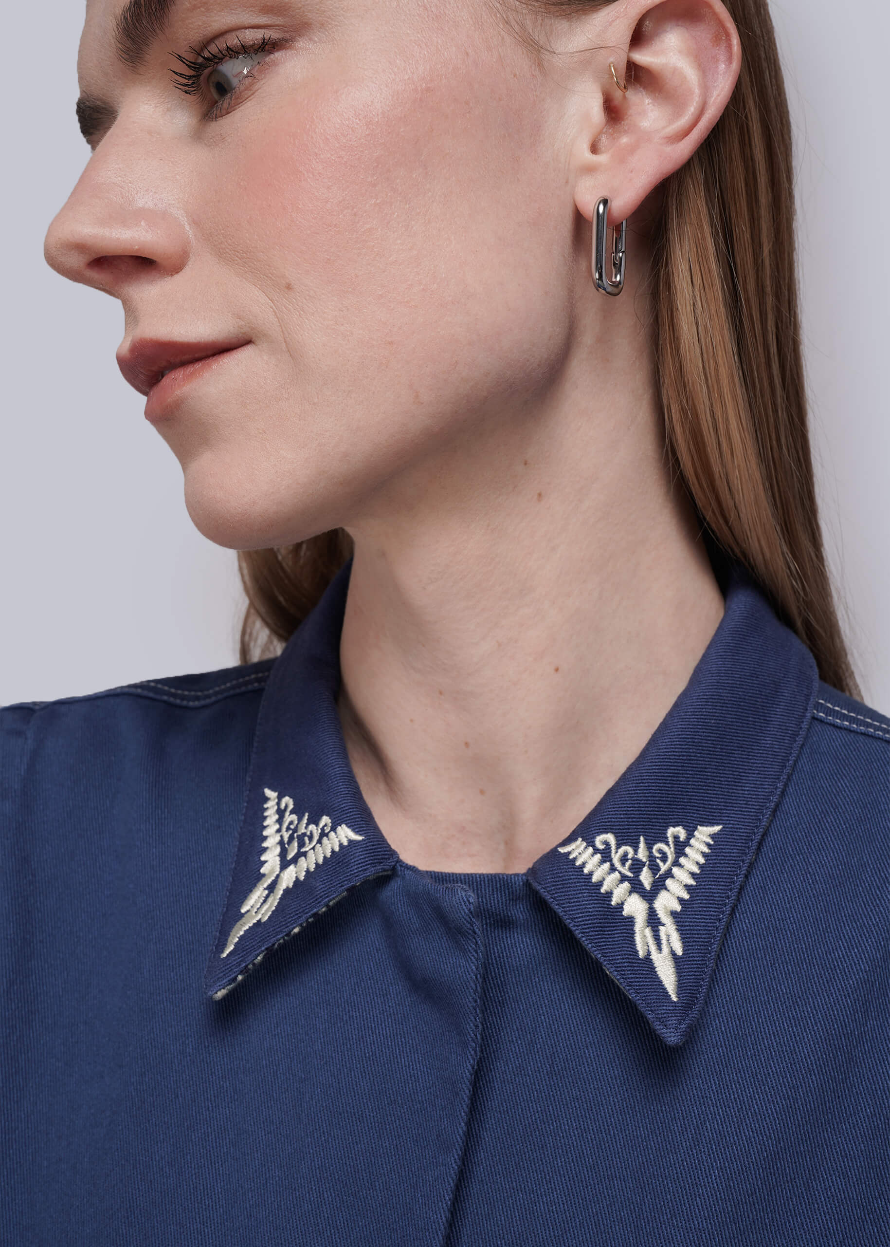 S=Close-up of a person wearing a navy blue coverall with white collar embroidery and hoop earrings.