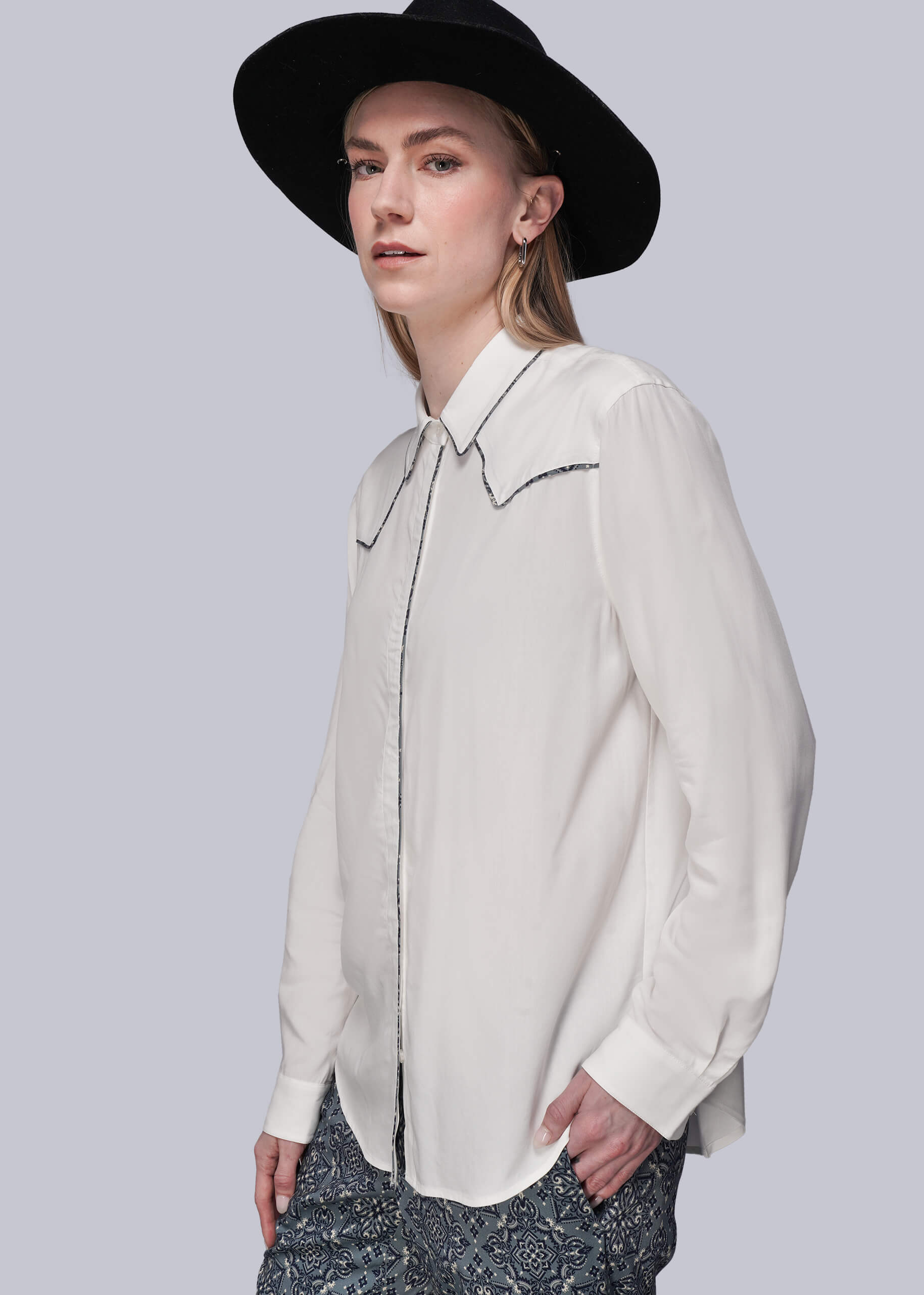 S=Woman wearing a white blouse with patterned trim and a black hat on a light gray background