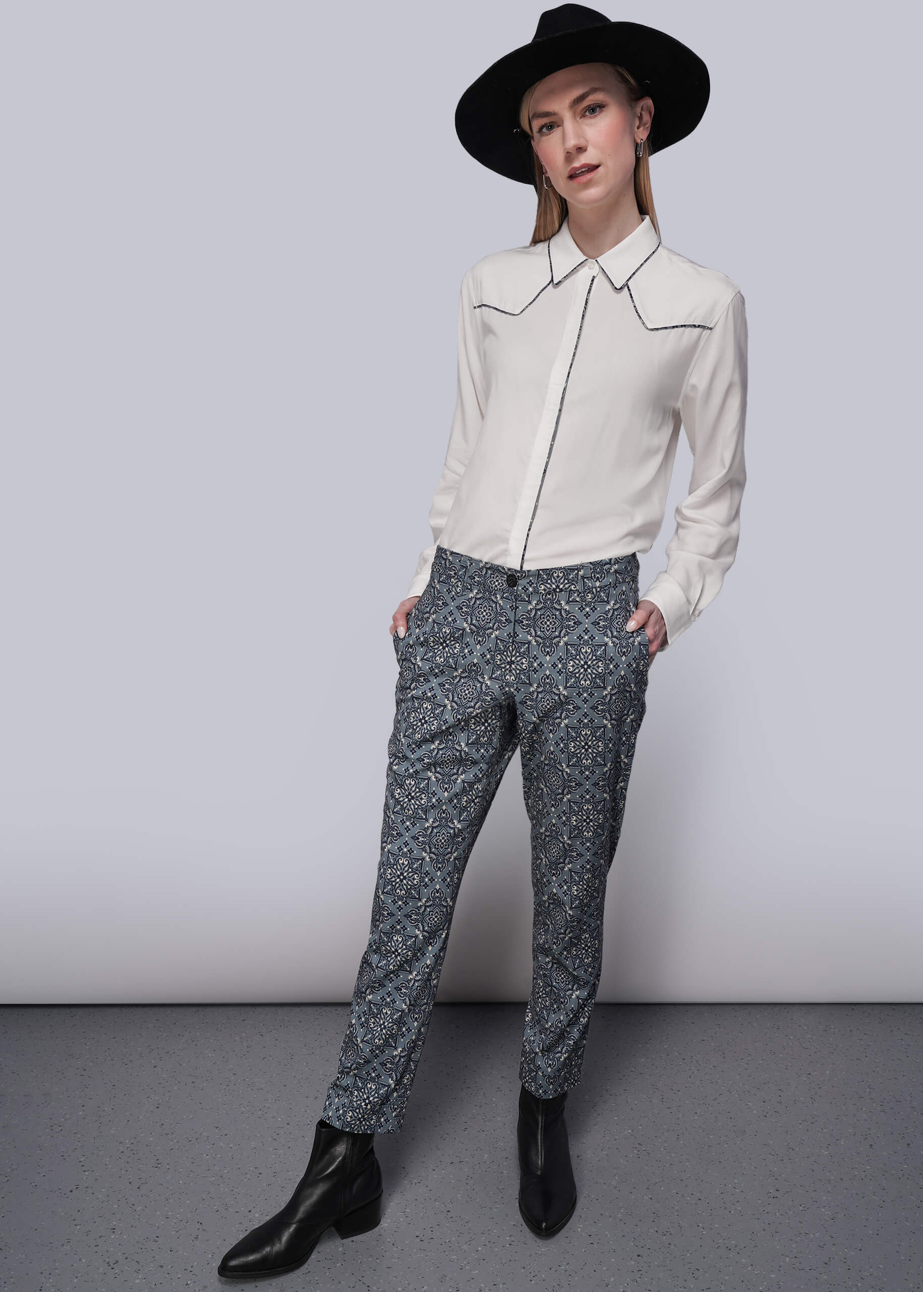 S=Person wearing a white blouse with decorative trim accents and patterned teal pants against a plain background