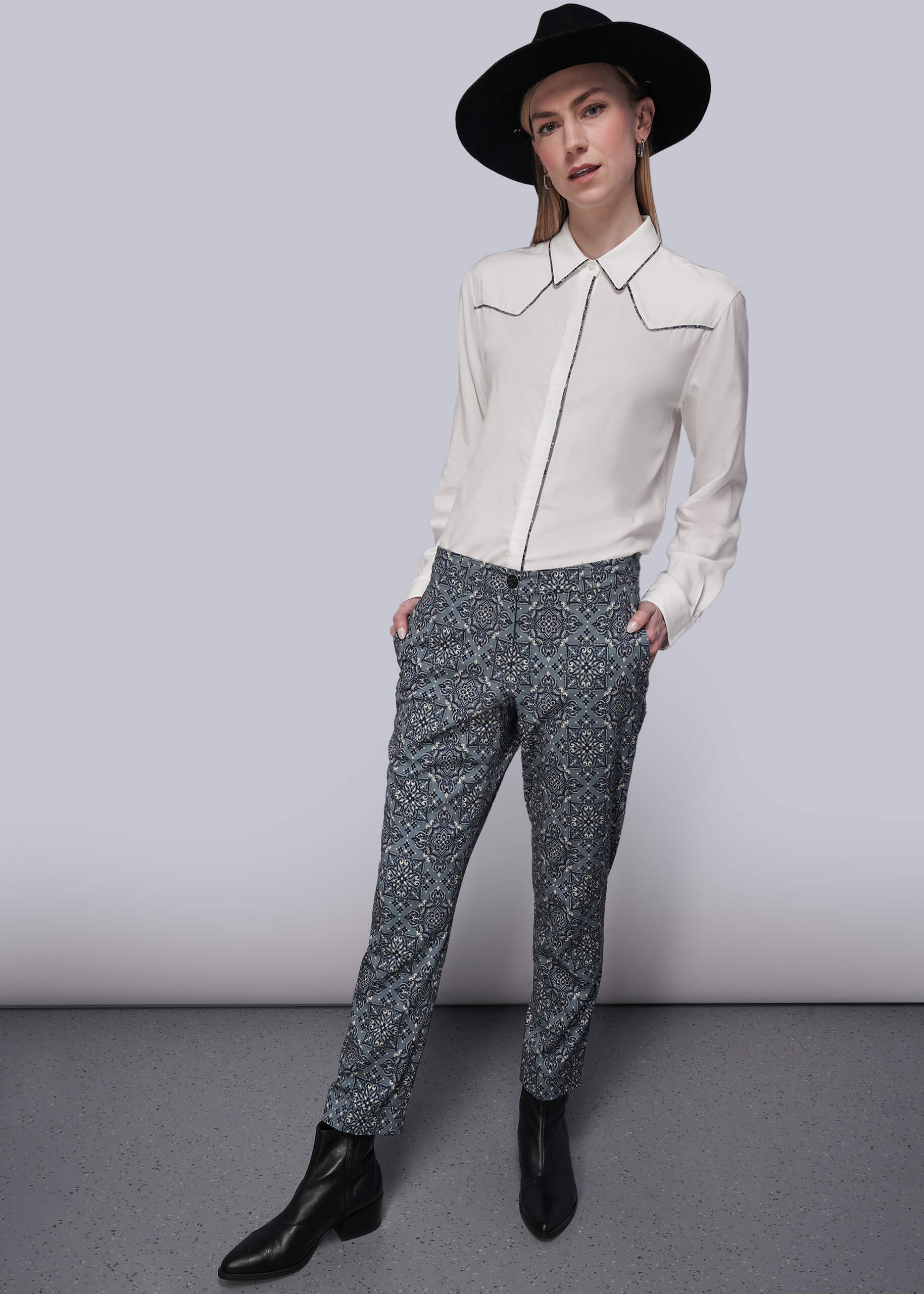 S=Person wearing a white blouse with patterned trim accents and patterned teal pants against a plain background