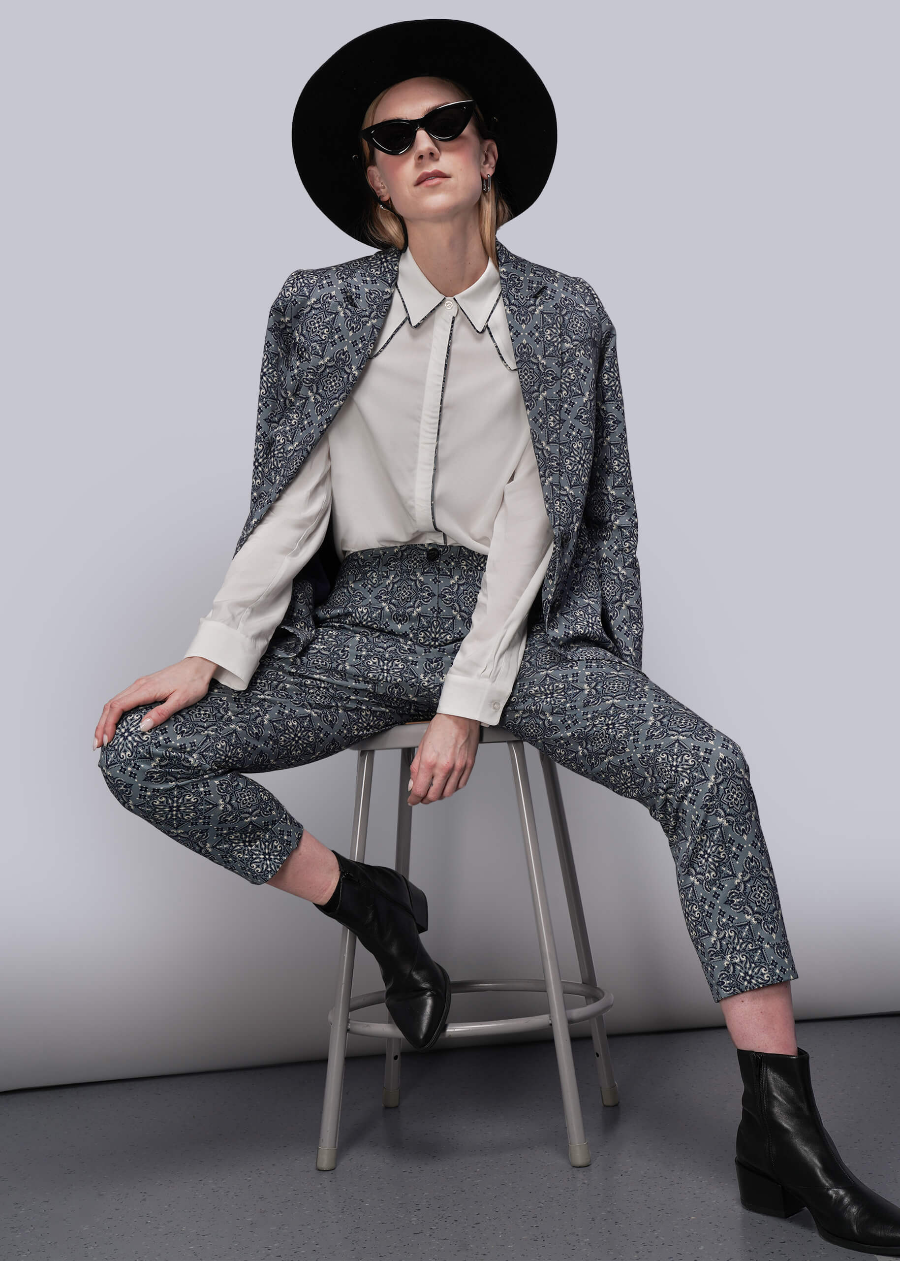 S=Person wearing a teal patterned suit and black hat sitting on a stool against a plain background