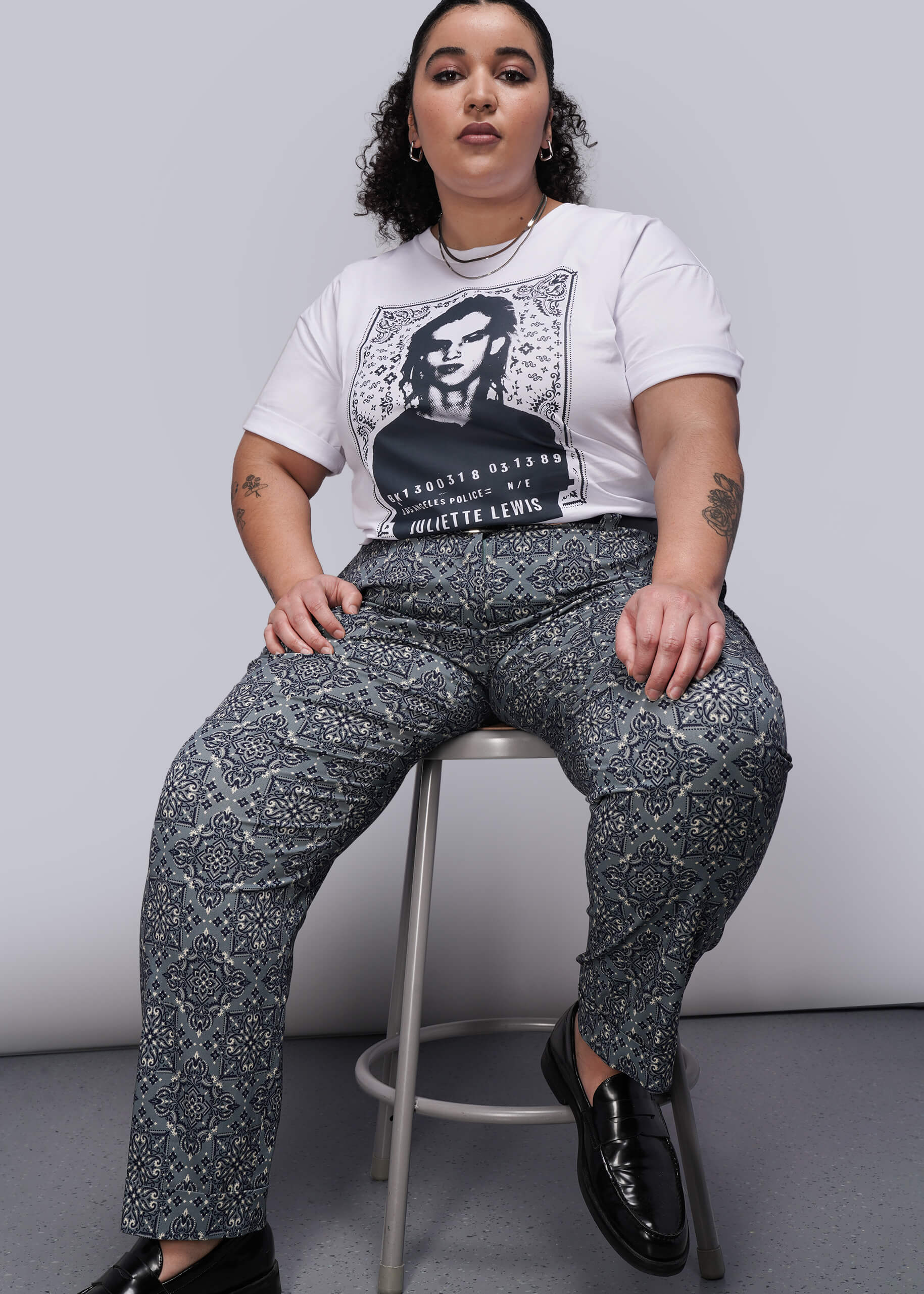 1X=Person sitting on a stool wearing a graphic t-shirt and patterned pants against a plain background